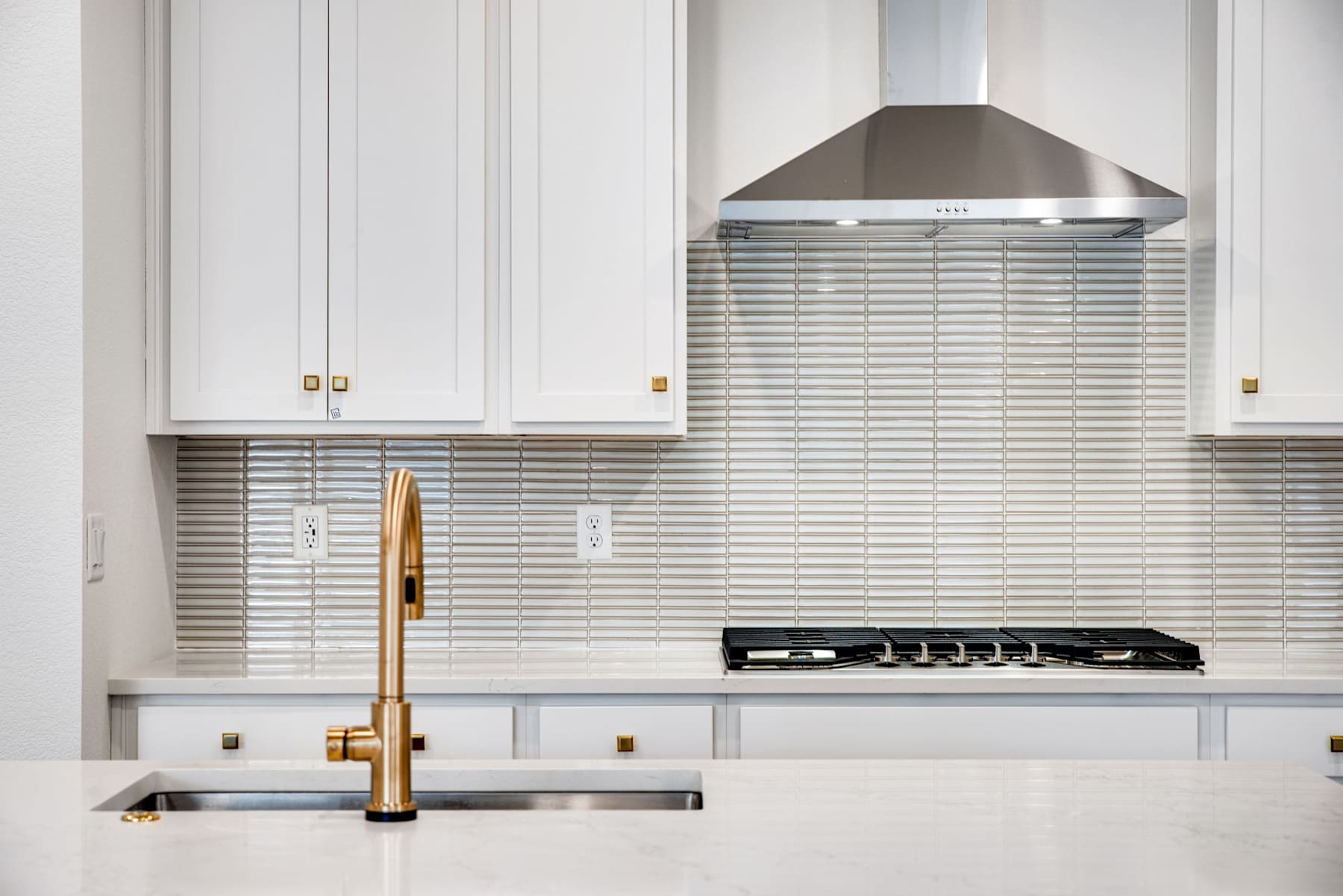 A modern, minimalist kitchen with white cabinets, a stainless steel range hood, and a gold-colored faucet over a stainless steel sink.