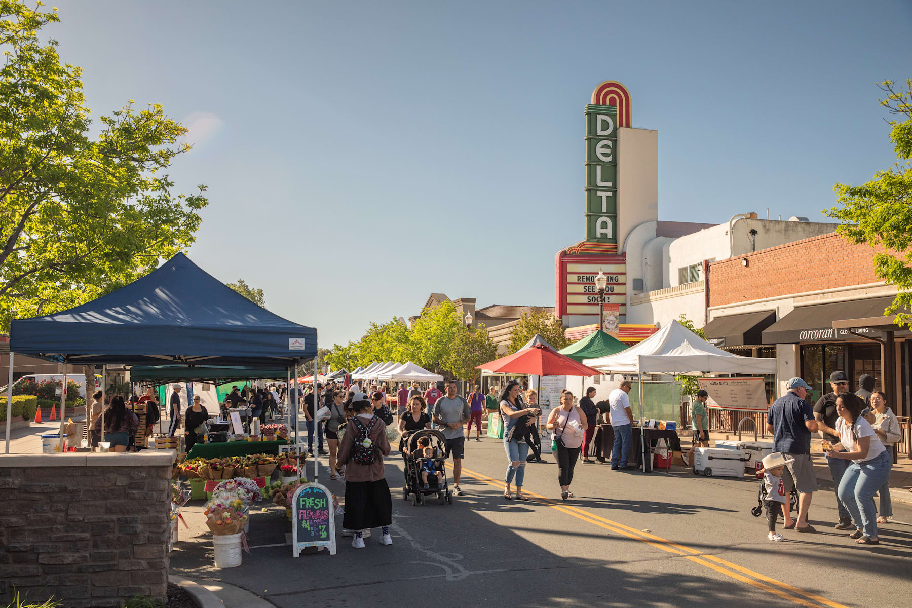A bustling outdoor market with various vendors and tents, set against the backdrop of a historic Delta theater building with its iconic neon sign.