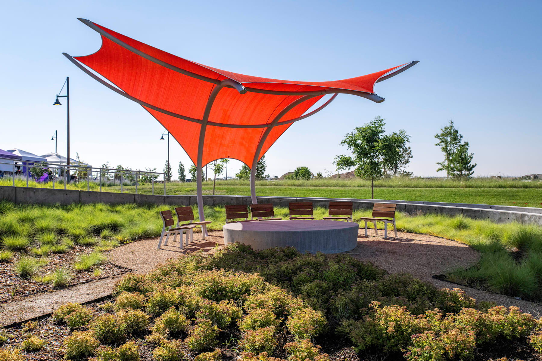 A large, vibrant red canopy structure stands in a landscaped outdoor area, surrounded by lush greenery and a paved seating area.