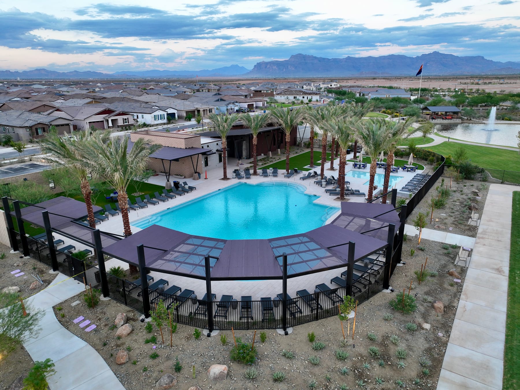 A large outdoor swimming pool surrounded by palm trees and buildings, with a mountainous landscape visible in the background.