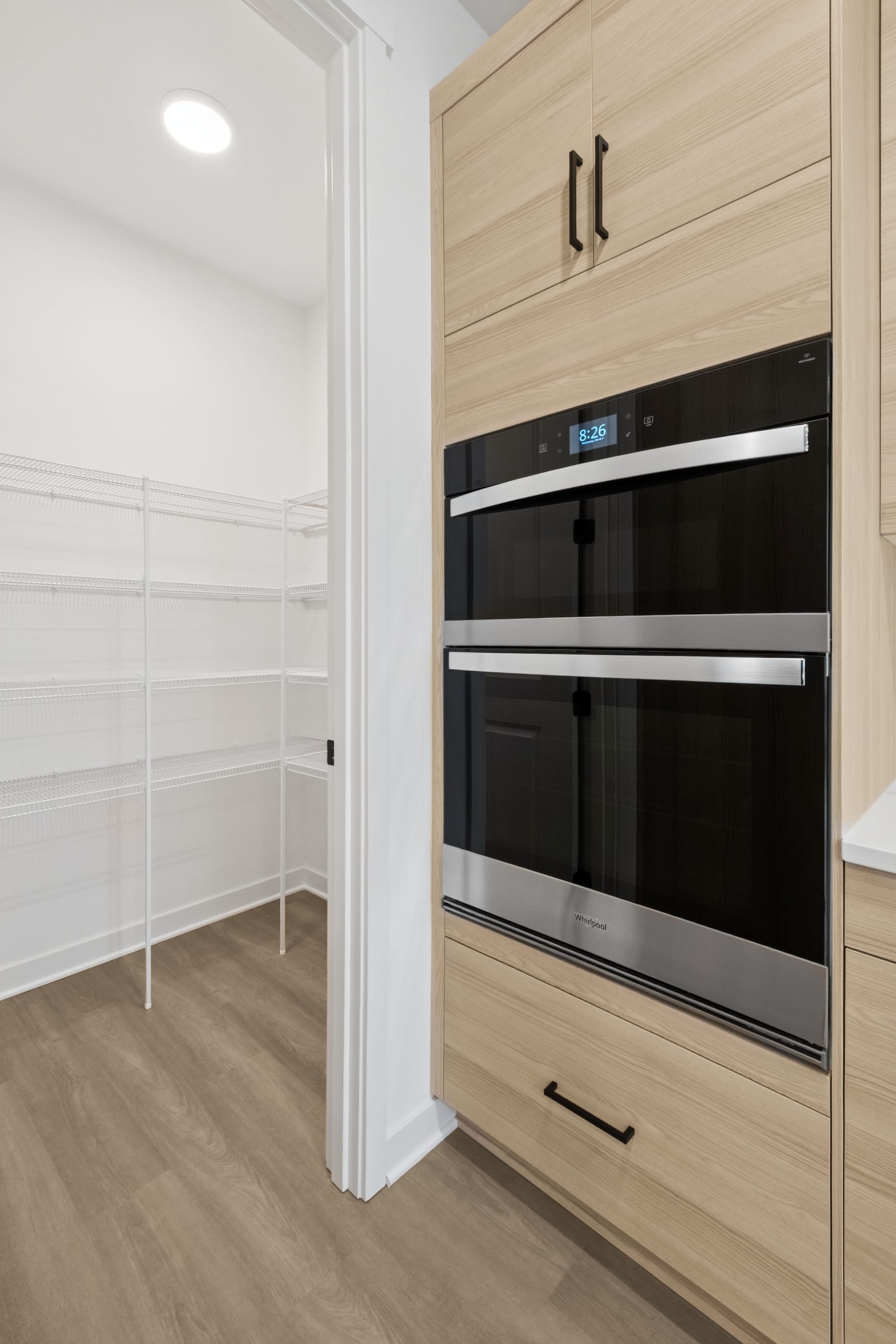 A modern kitchen with light-colored wood cabinets, a built-in oven, and a partially visible walk-in pantry in the background.