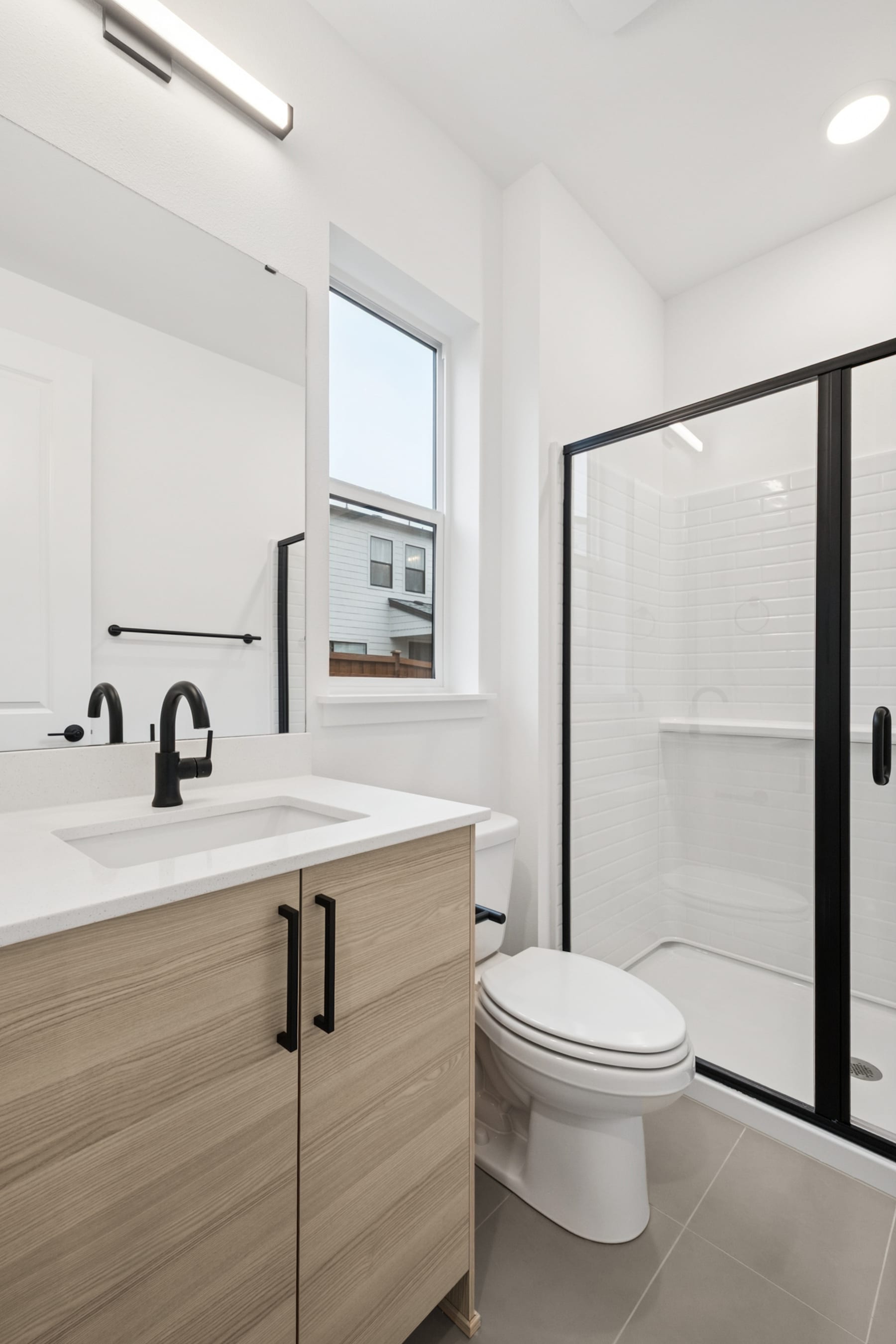 A modern and minimalist bathroom with a wooden vanity, white countertop, and a large glass shower enclosure, set against a bright and airy backdrop.
