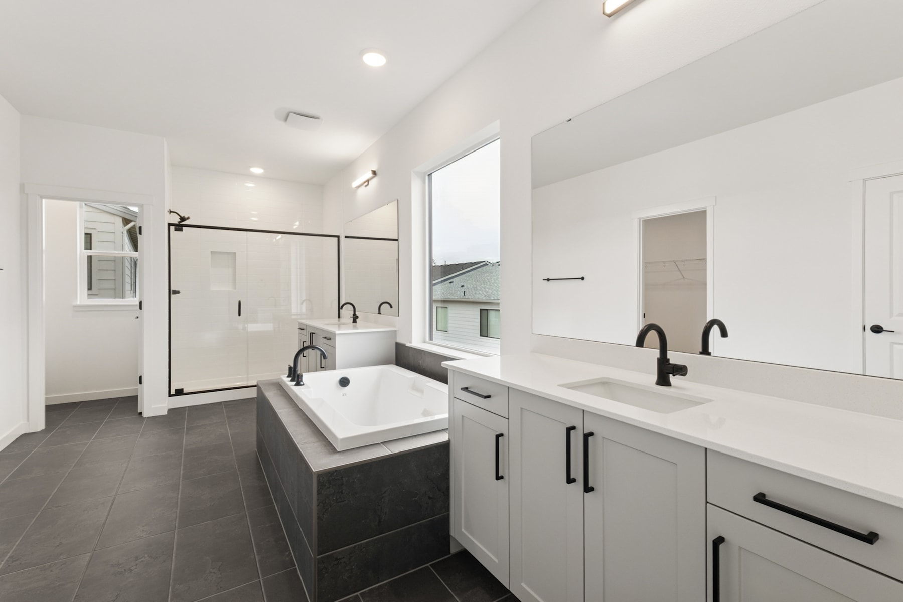 A modern, minimalist bathroom with a large bathtub, double vanity, and dark tile flooring, set against a bright, airy backdrop.