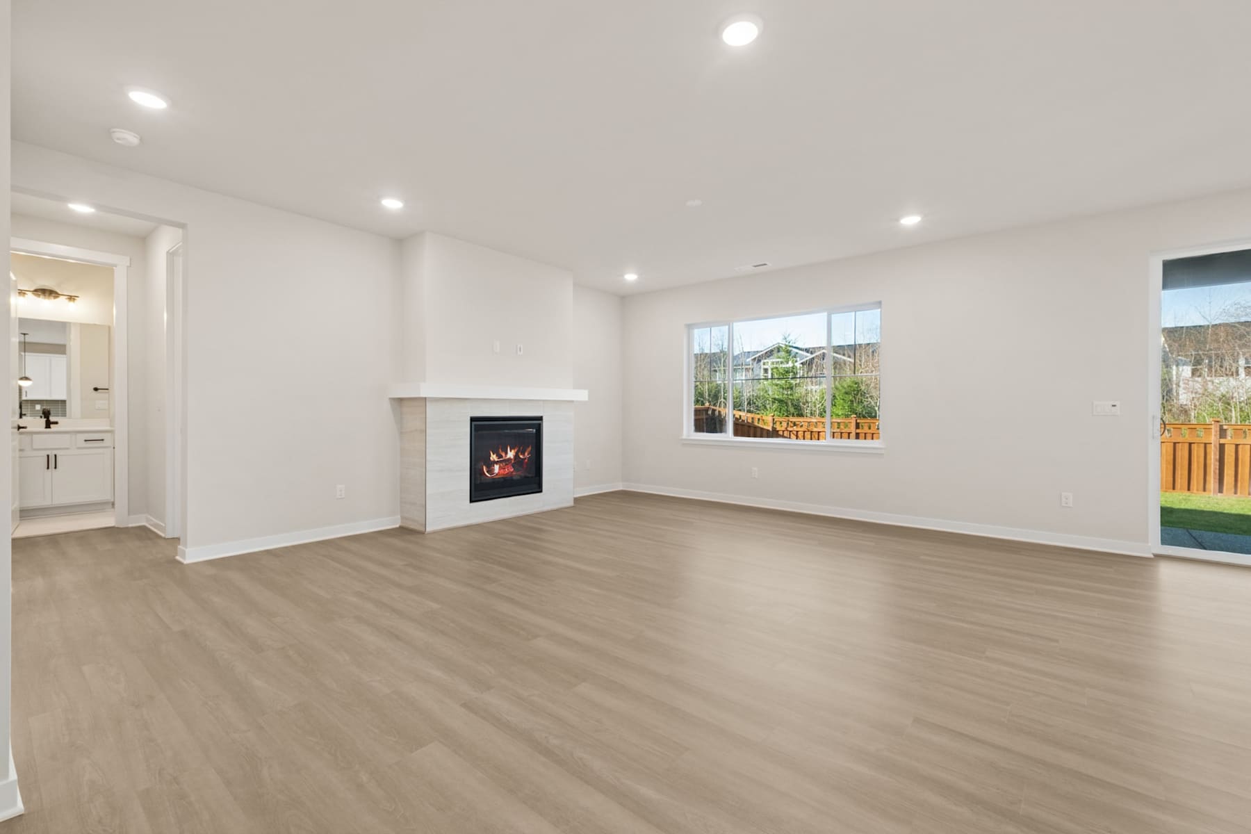 A spacious, well-lit room with a fireplace, hardwood floors, and large windows overlooking a backyard.