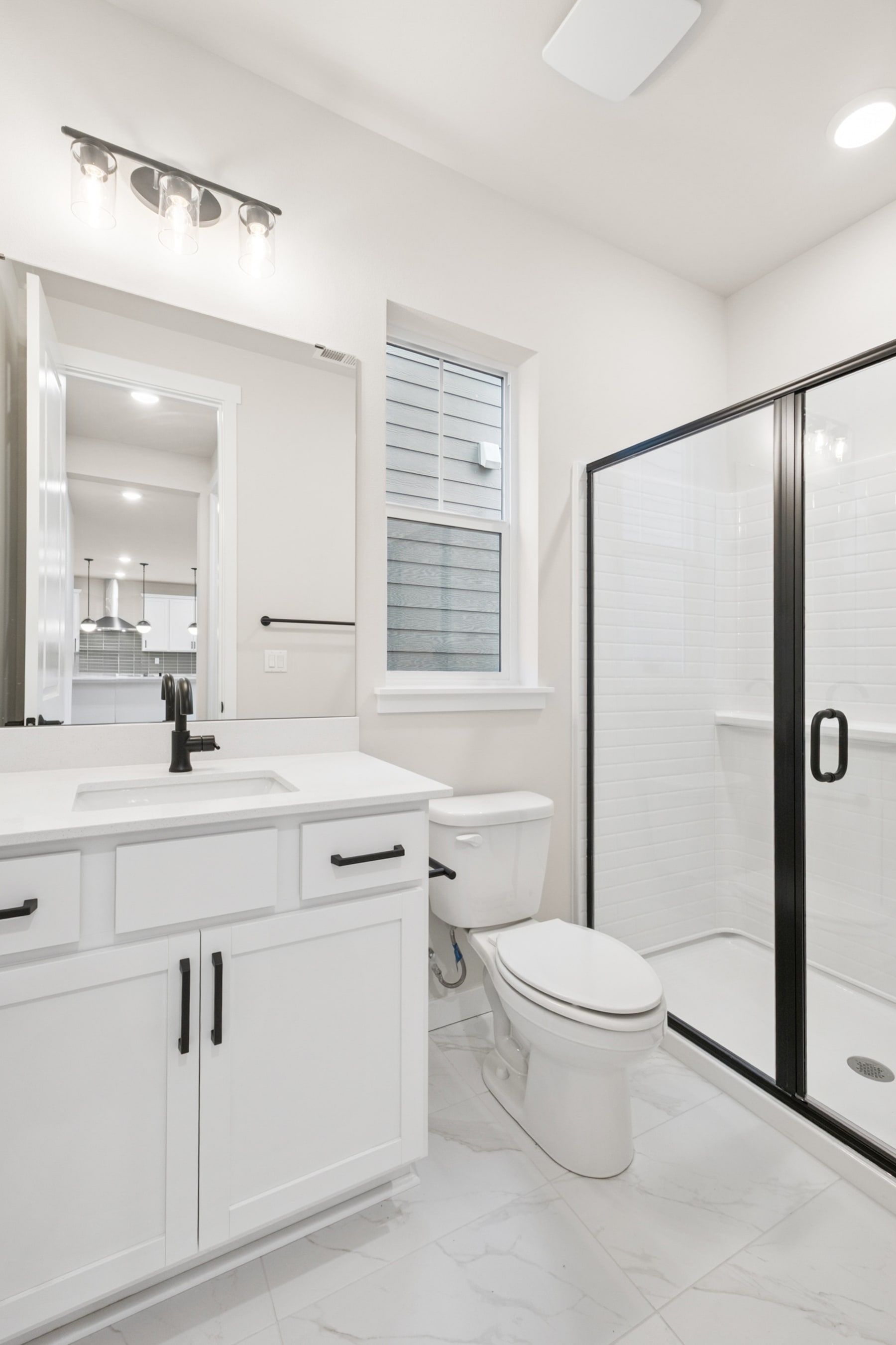 A modern, well-lit bathroom with white cabinets, a vanity, and a toilet, along with a large mirror and a window providing natural light.