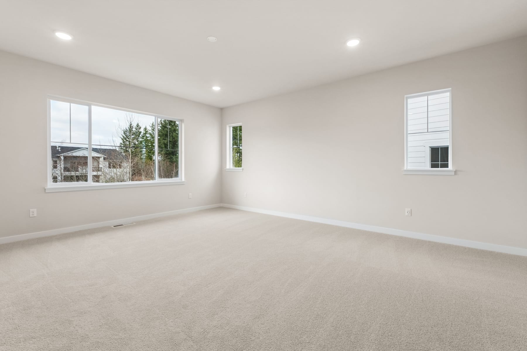 A spacious, well-lit room with white walls, plush carpeting, and large windows overlooking a residential neighborhood.