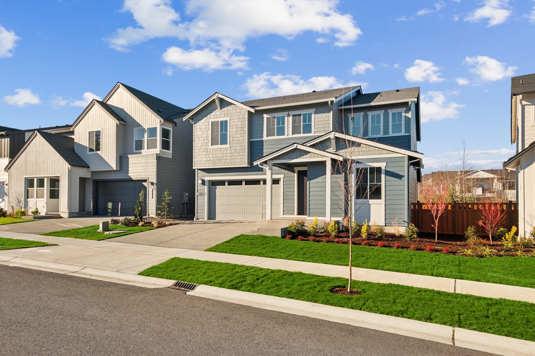 A residential neighborhood with modern, two-story houses featuring a well-manicured lawn and landscaping in the foreground, set against a backdrop of a blue sky with fluffy white clouds.