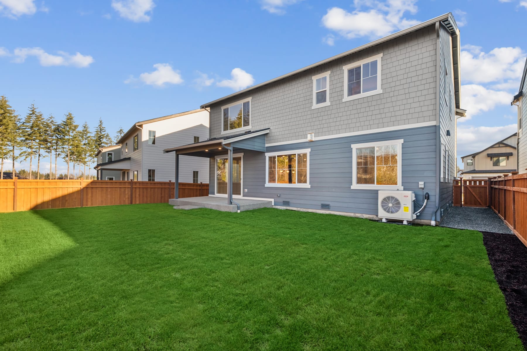A well-maintained residential property with a lush green lawn in the foreground and a two-story gray house with a porch and windows in the background, surrounded by trees and a wooden fence.