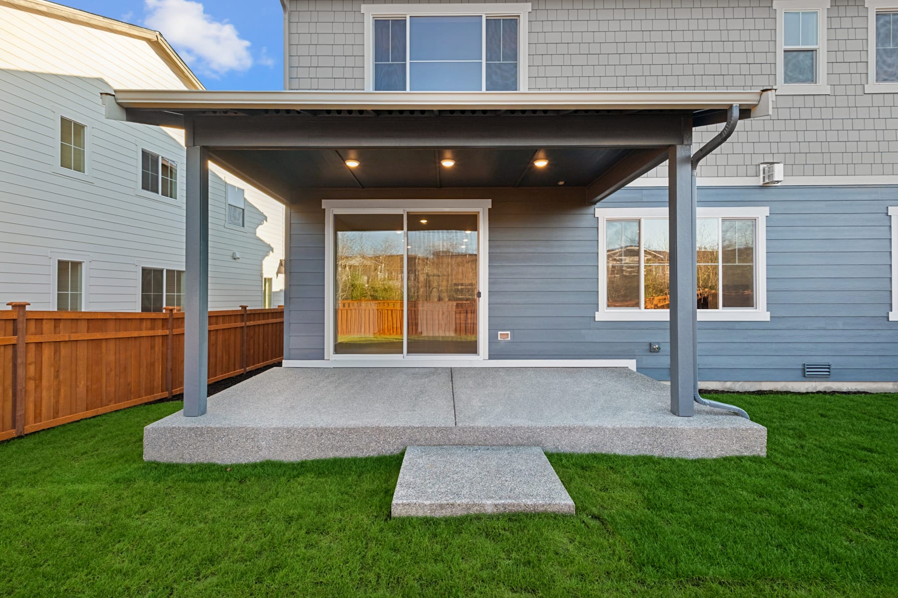 A modern two-story house with a covered porch, surrounded by a well-manicured lawn and a wooden fence in the background.