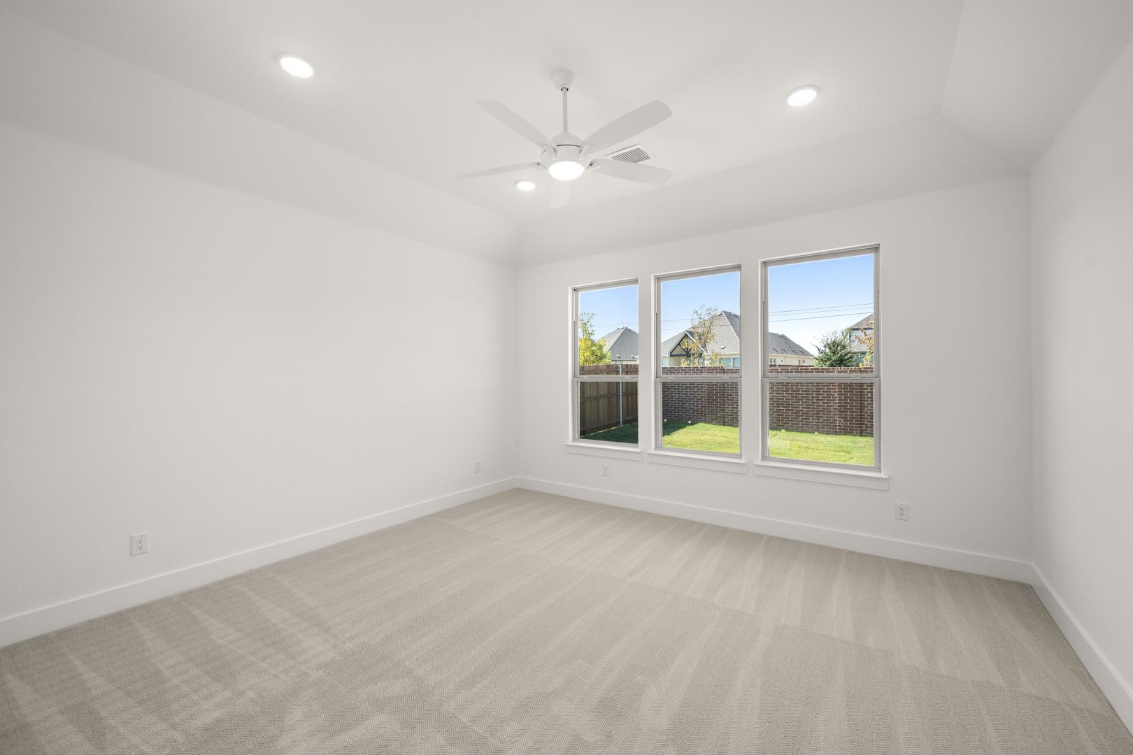 A bright, spacious room with large windows overlooking a backyard, featuring a ceiling fan and hardwood flooring.