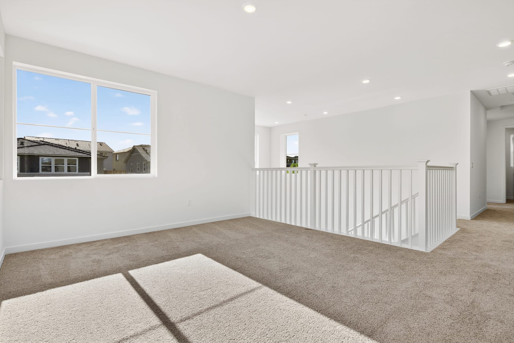 A bright and spacious interior with a carpeted floor, white walls, and a railing leading to an upper level. The room appears to be a hallway or entryway with a large window providing natural light.