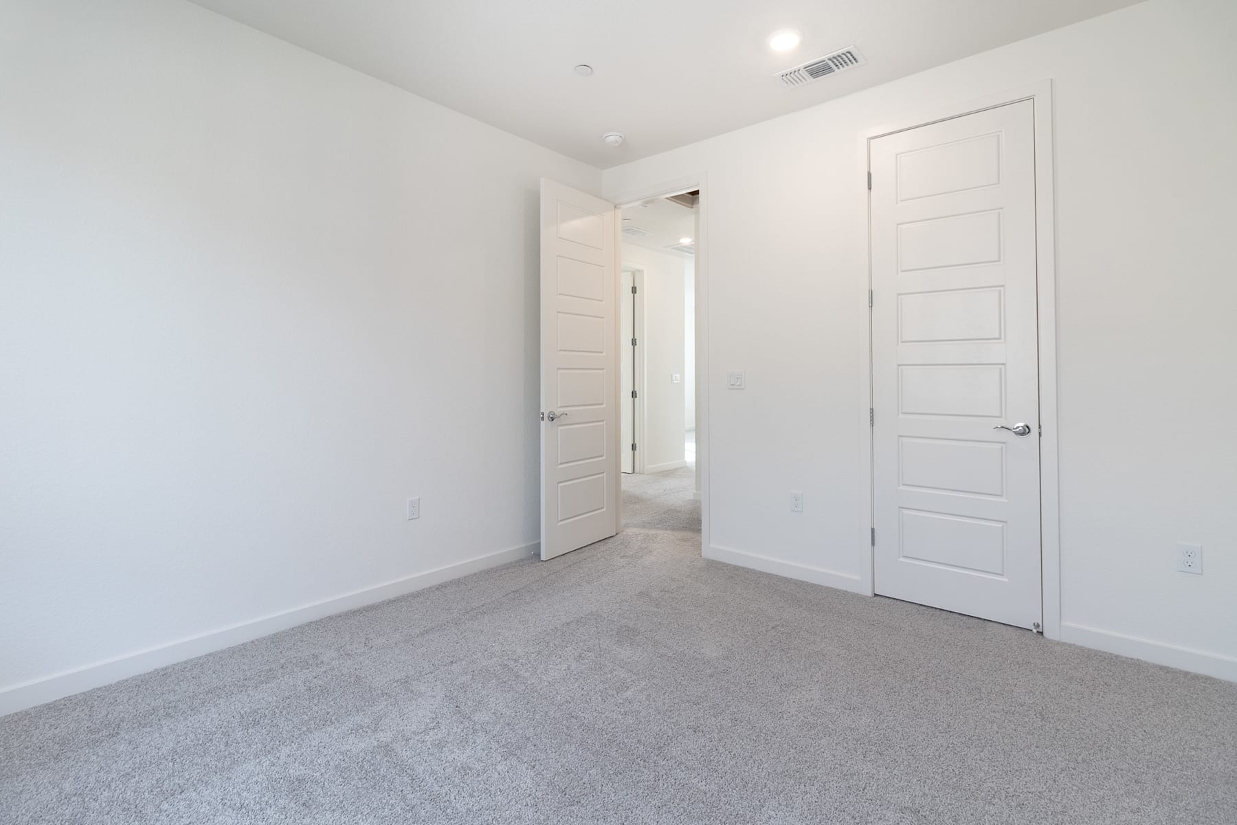A spacious, minimalist bedroom with white walls, a carpeted floor, and a closed door leading to another room.