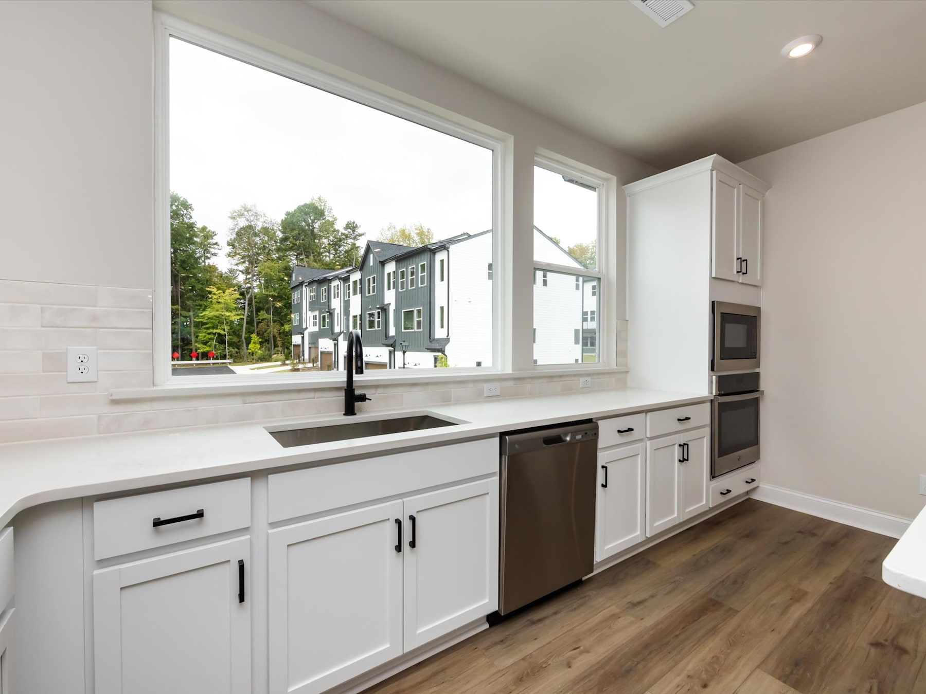 A modern, well-lit kitchen with white cabinets, stainless steel appliances, and large windows overlooking a wooded outdoor area.
