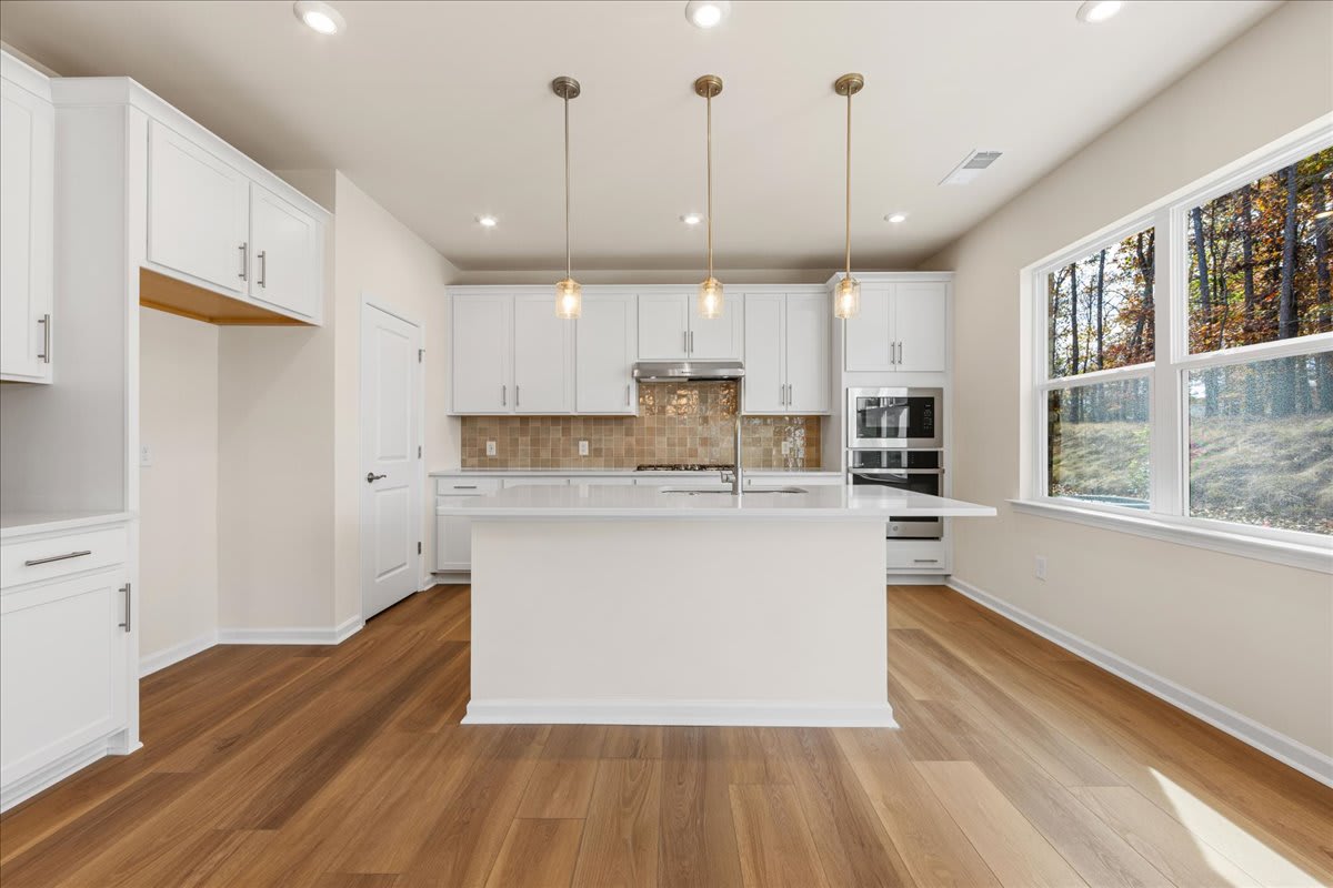 A modern, bright kitchen with white cabinets, wooden floors, and large windows overlooking a wooded area.
