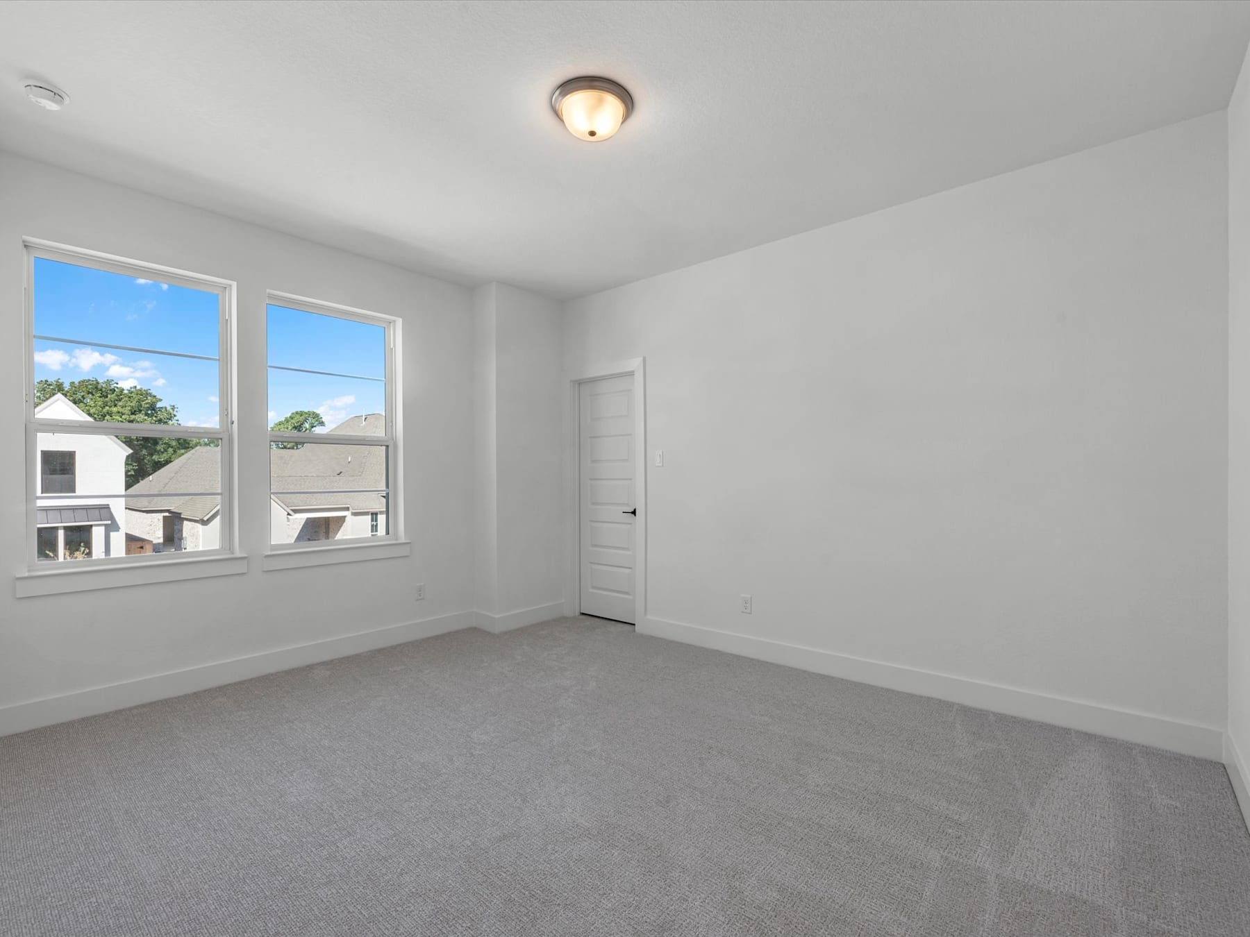 A bright, empty room with white walls, a ceiling light fixture, and large windows overlooking a residential neighborhood.
