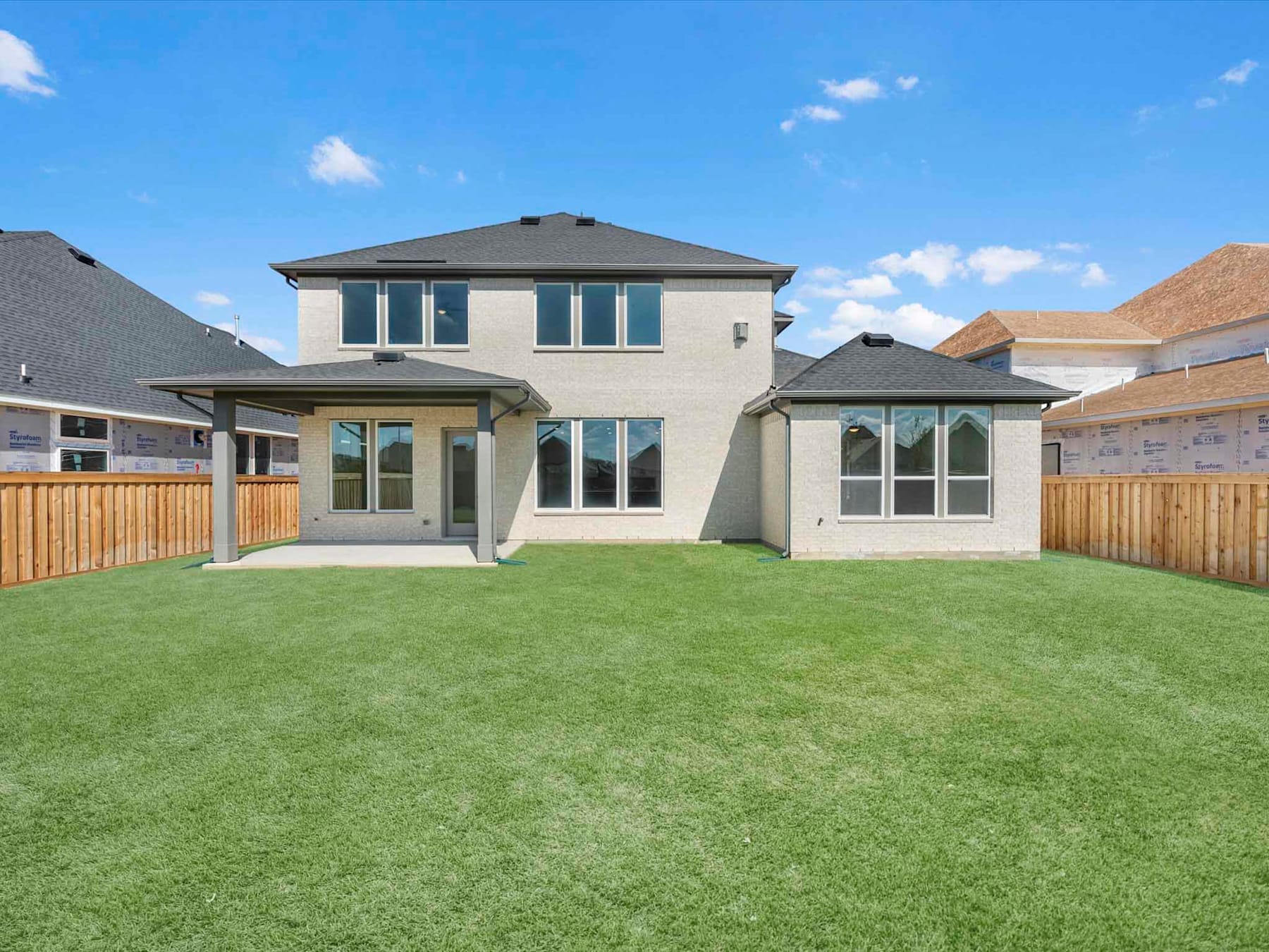 A modern two-story house with a large grassy yard and a wooden fence in the foreground, set against a clear blue sky with scattered clouds.