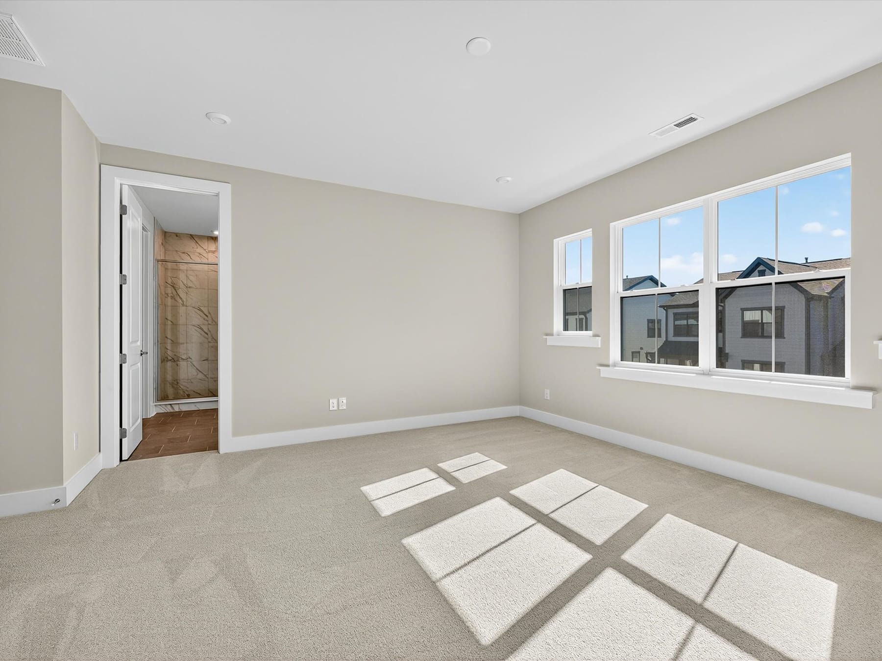 A spacious, well-lit room with a neutral color scheme, featuring a carpeted floor and large windows overlooking the exterior.