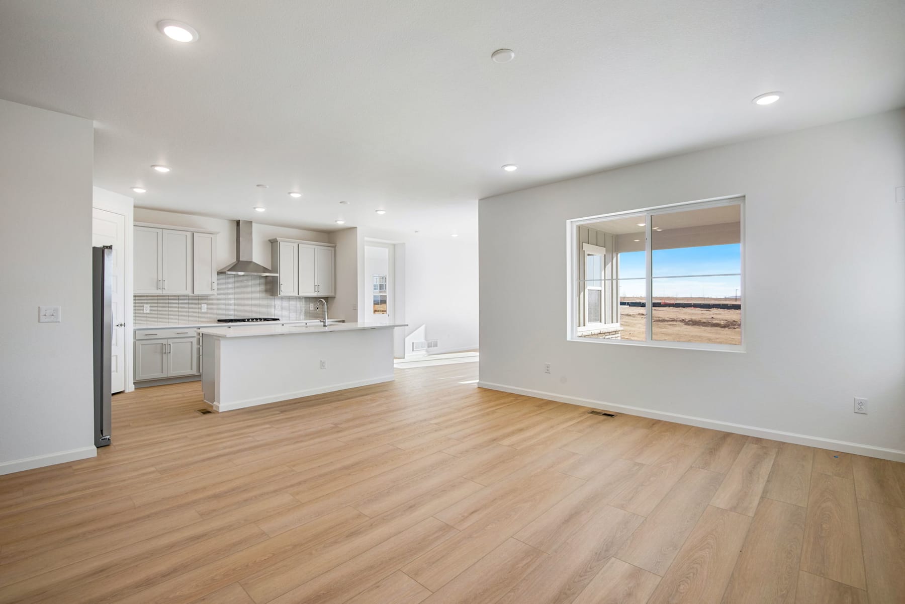 A spacious, modern kitchen with white cabinets and appliances opens up to a bright, airy living room with hardwood floors and a large window overlooking a scenic outdoor landscape.