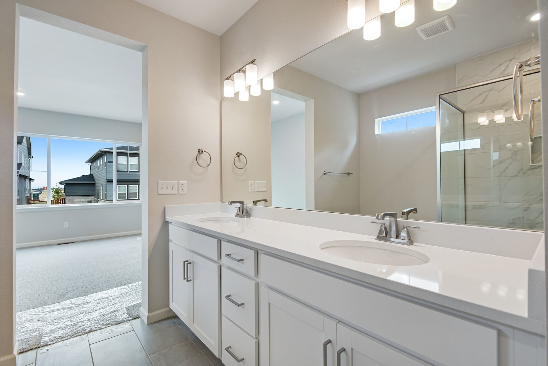 A modern, well-lit bathroom with a double vanity, a large mirror, and a view of the exterior through a window.
