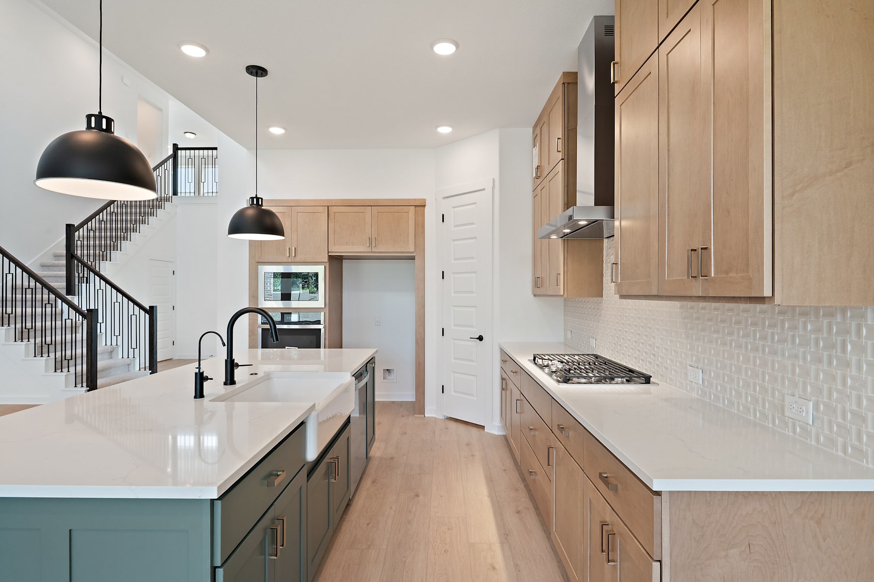 A modern, open-concept kitchen with light wood cabinets, a white countertop, and black pendant lights, leading into a hallway with a staircase.