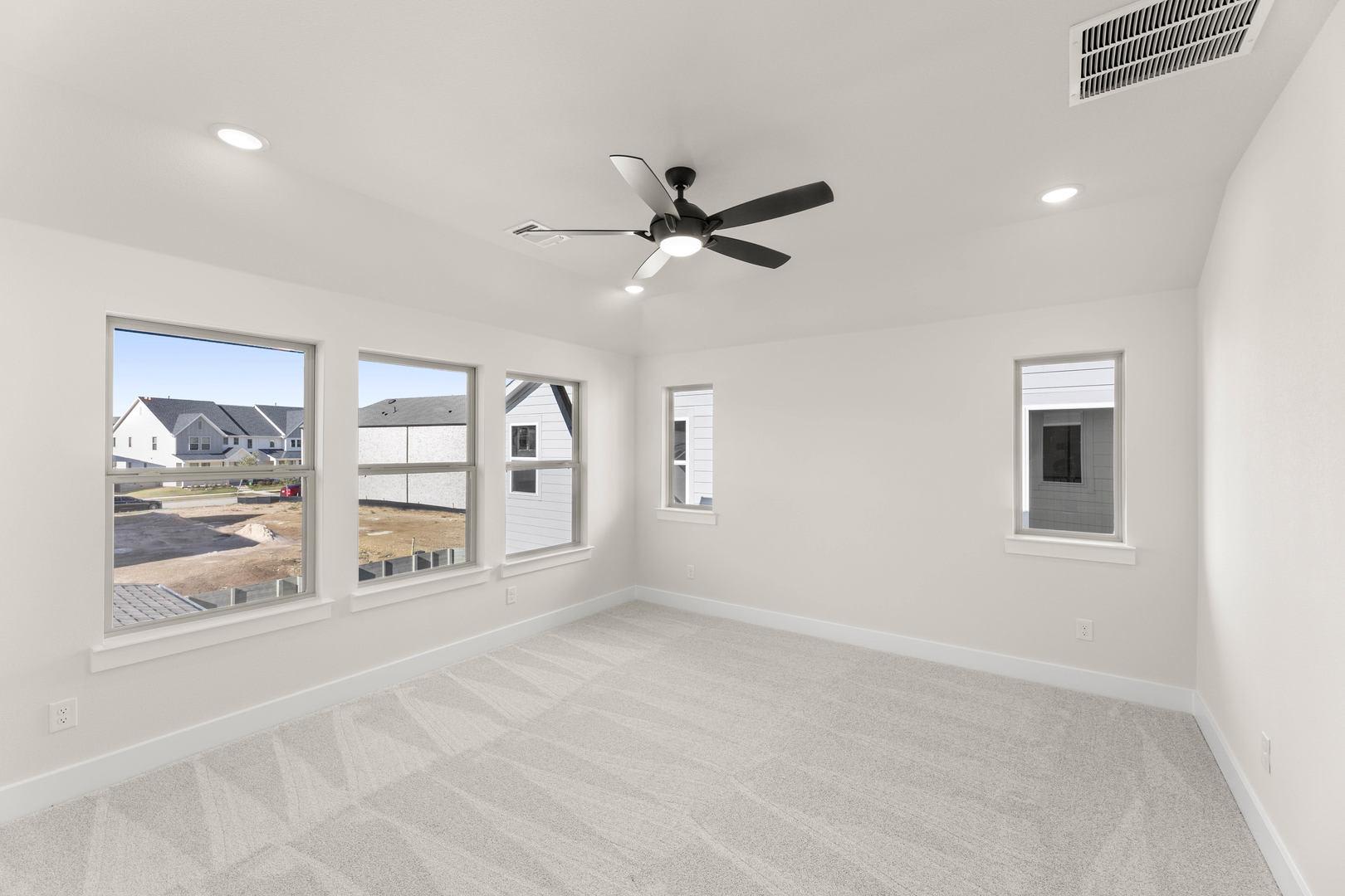 A spacious, well-lit room with a ceiling fan, large windows, and a hardwood floor, overlooking a residential neighborhood in the background.
