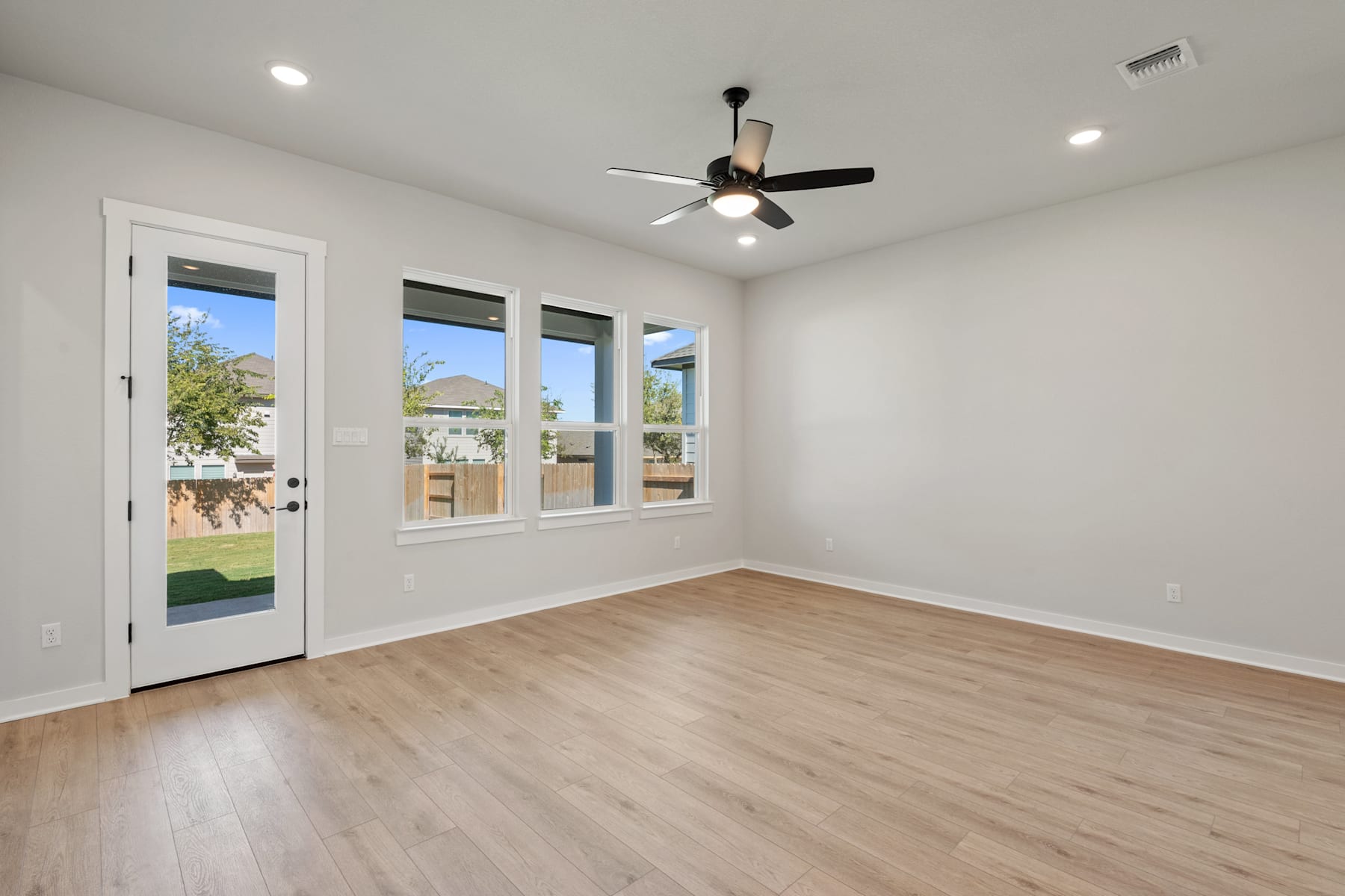 A bright, spacious room with a ceiling fan, large windows, and a wooden floor.