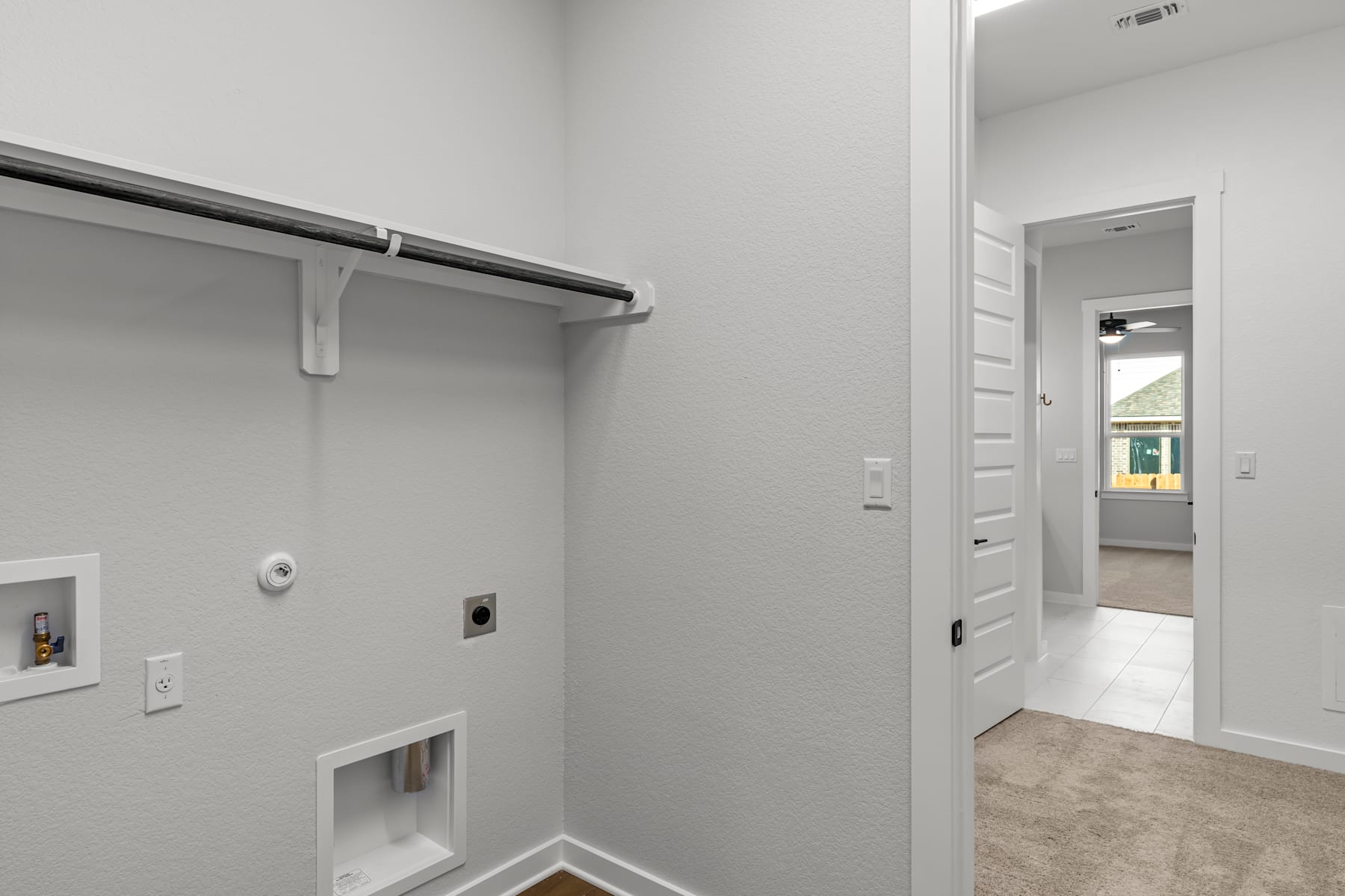 A hallway with a white wall, a closet rod, and a doorway leading to another room.