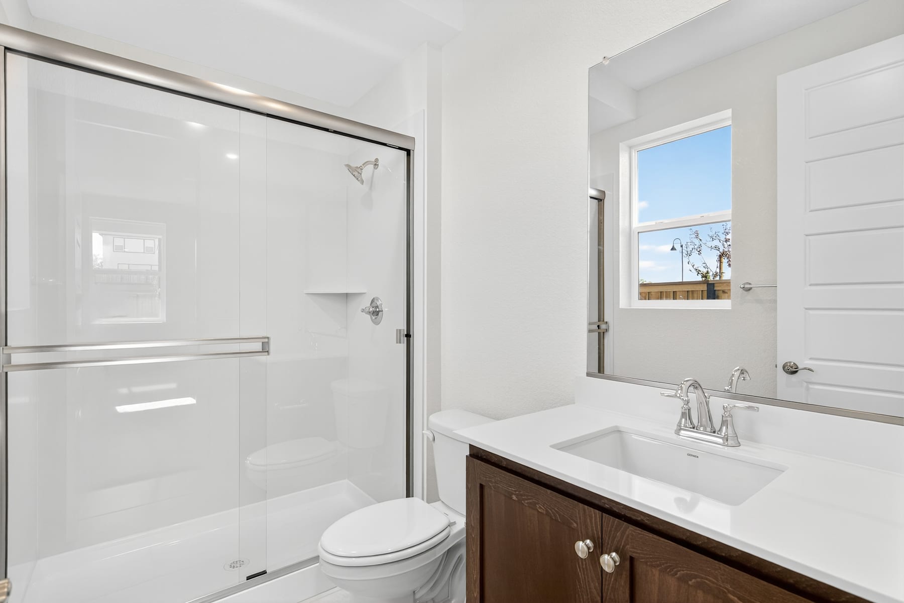 A modern, well-lit bathroom with a glass-enclosed shower, a vanity with a sink, and a toilet. The room has a clean, minimalist design with white walls and a wooden vanity.