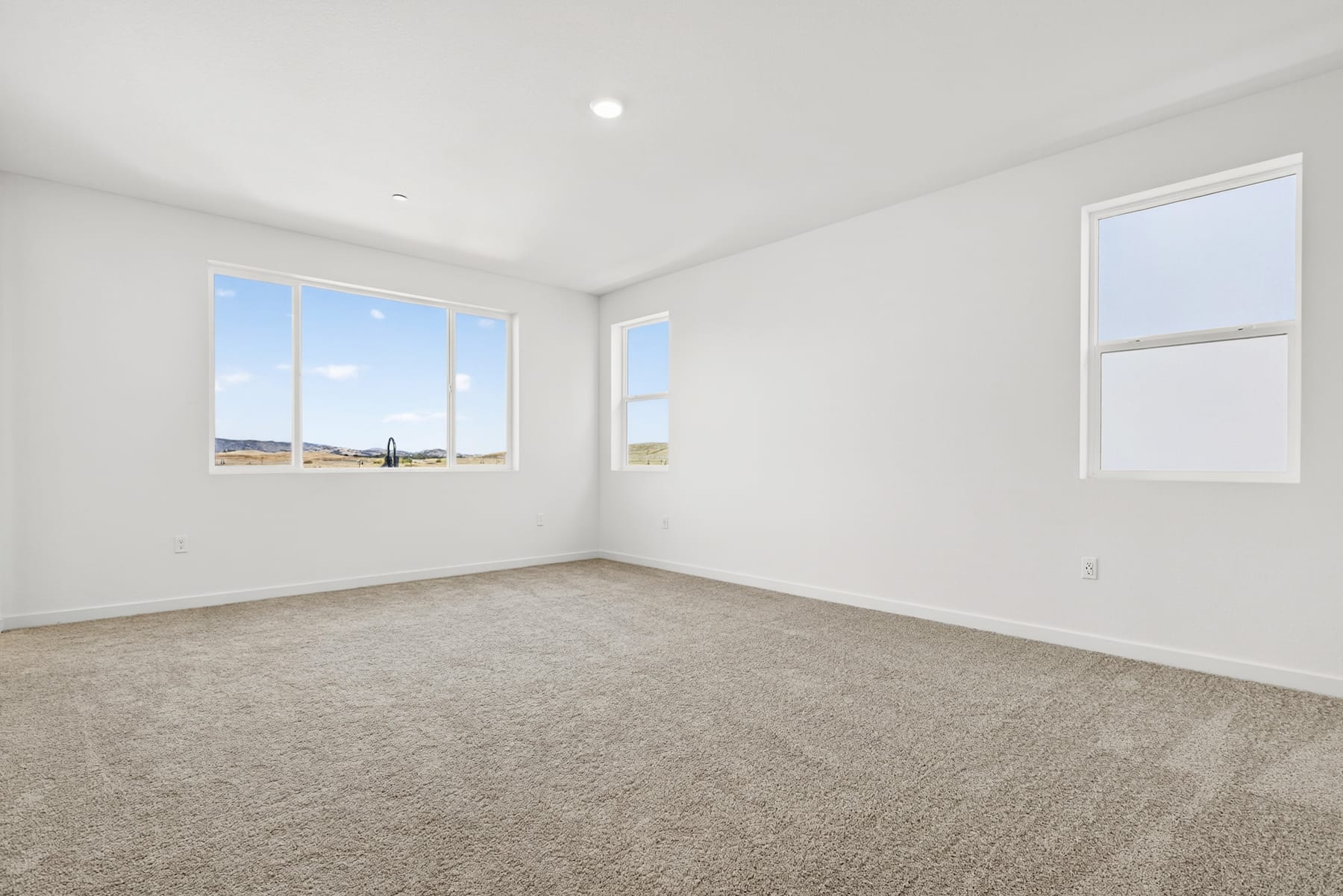 A spacious, well-lit room with large windows overlooking a scenic outdoor landscape, featuring a carpeted floor and bare walls, creating a clean and minimalist atmosphere.
