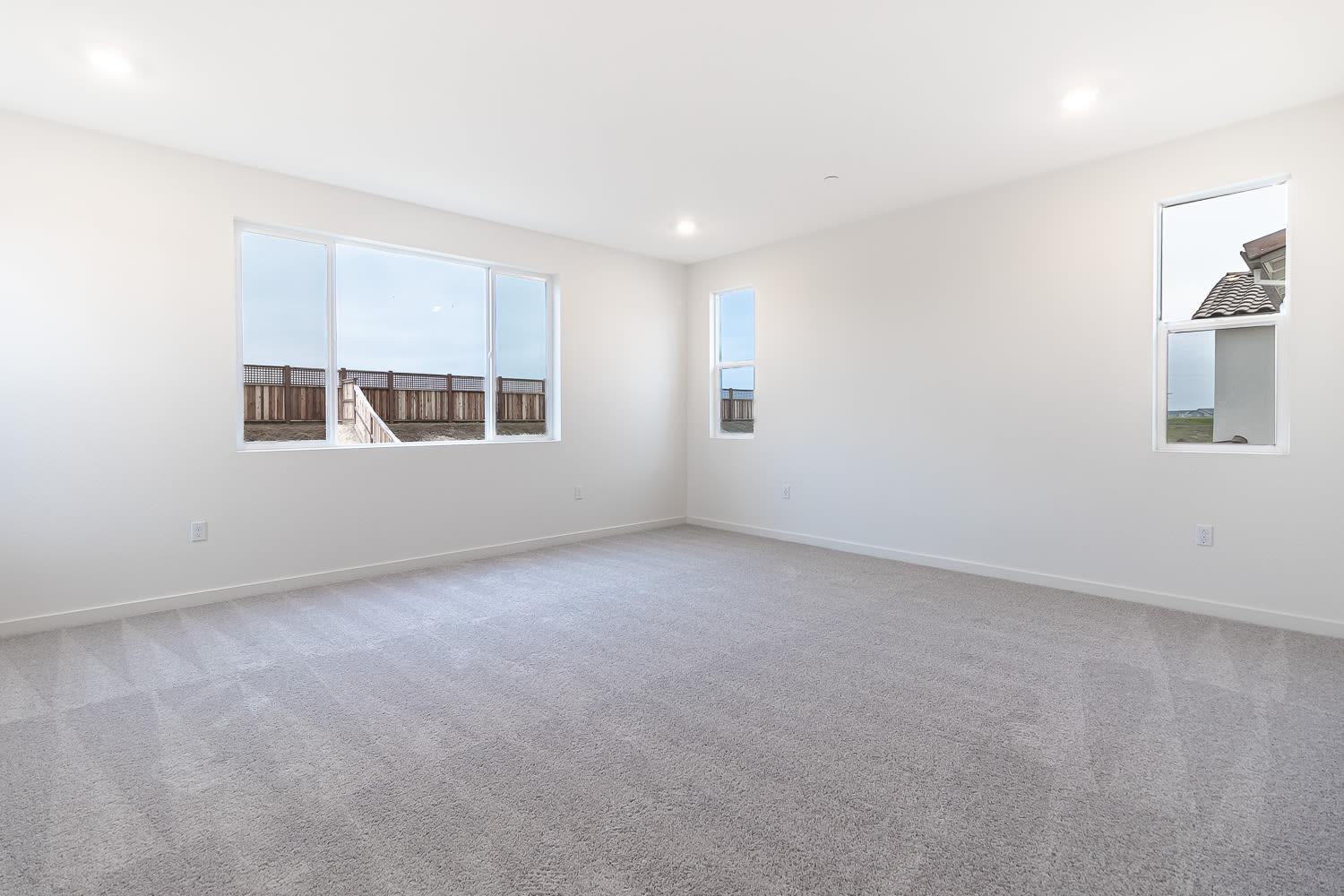 A spacious, well-lit room with white walls, hardwood floors, and large windows providing a view of the exterior.