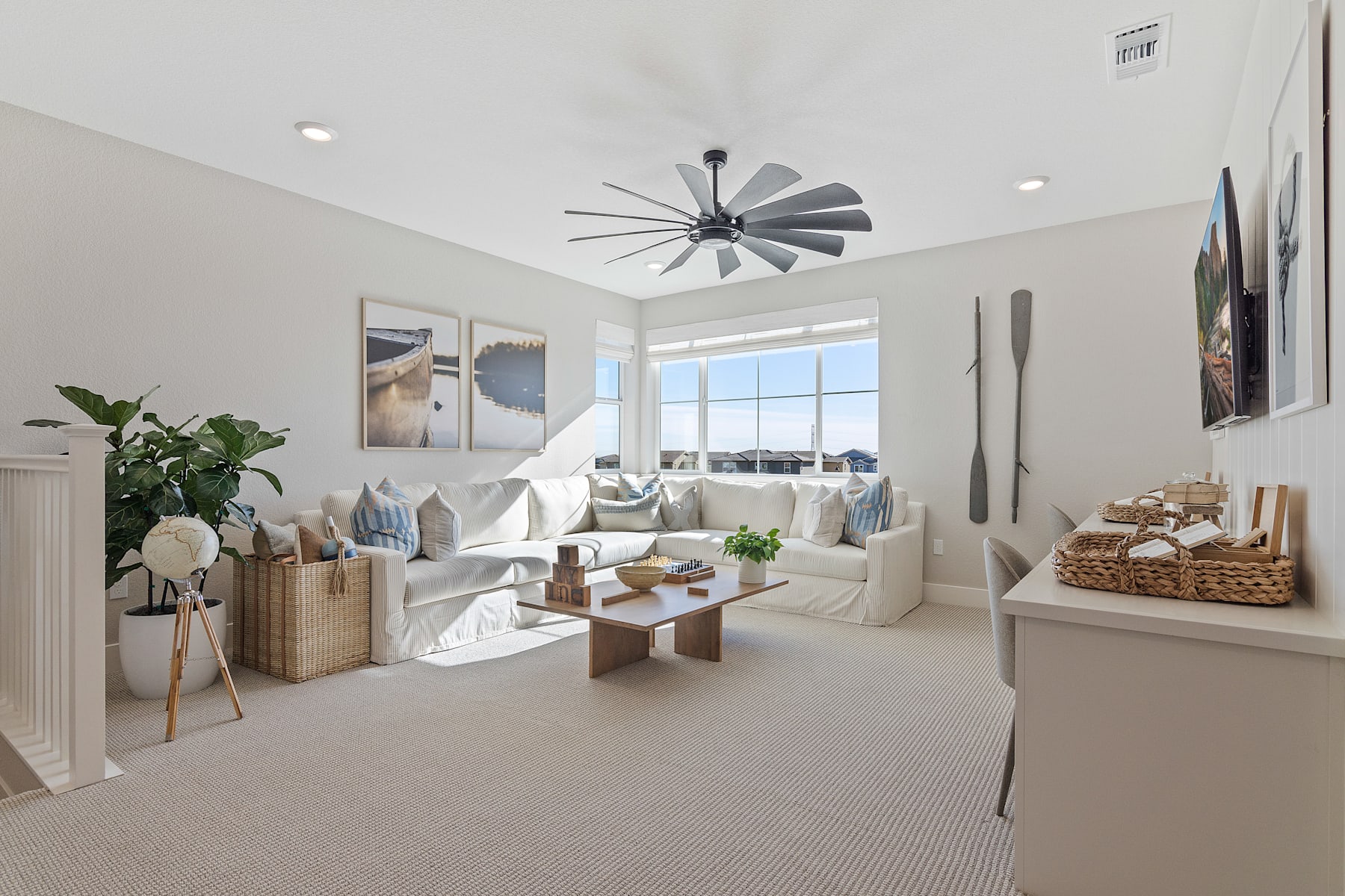 A cozy and bright living room with a large window, a modern ceiling fan, and various decorative elements such as plants, baskets, and artwork.