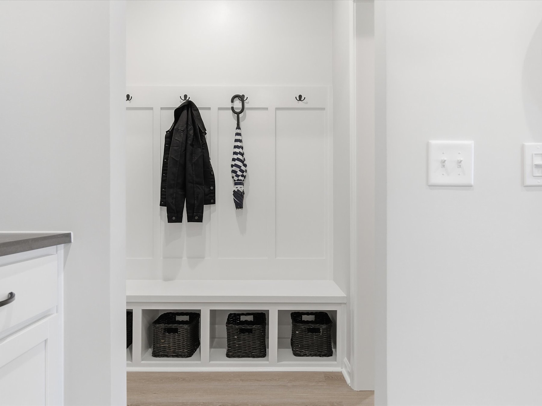 A simple, minimalist entryway with a white wall, a row of hooks for hanging coats or bags, and three storage baskets below.