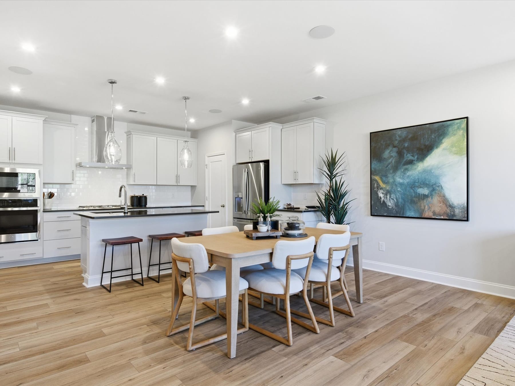 A modern, open-concept kitchen and dining area with white cabinets, stainless steel appliances, a wooden dining table, and a large abstract painting on the wall.