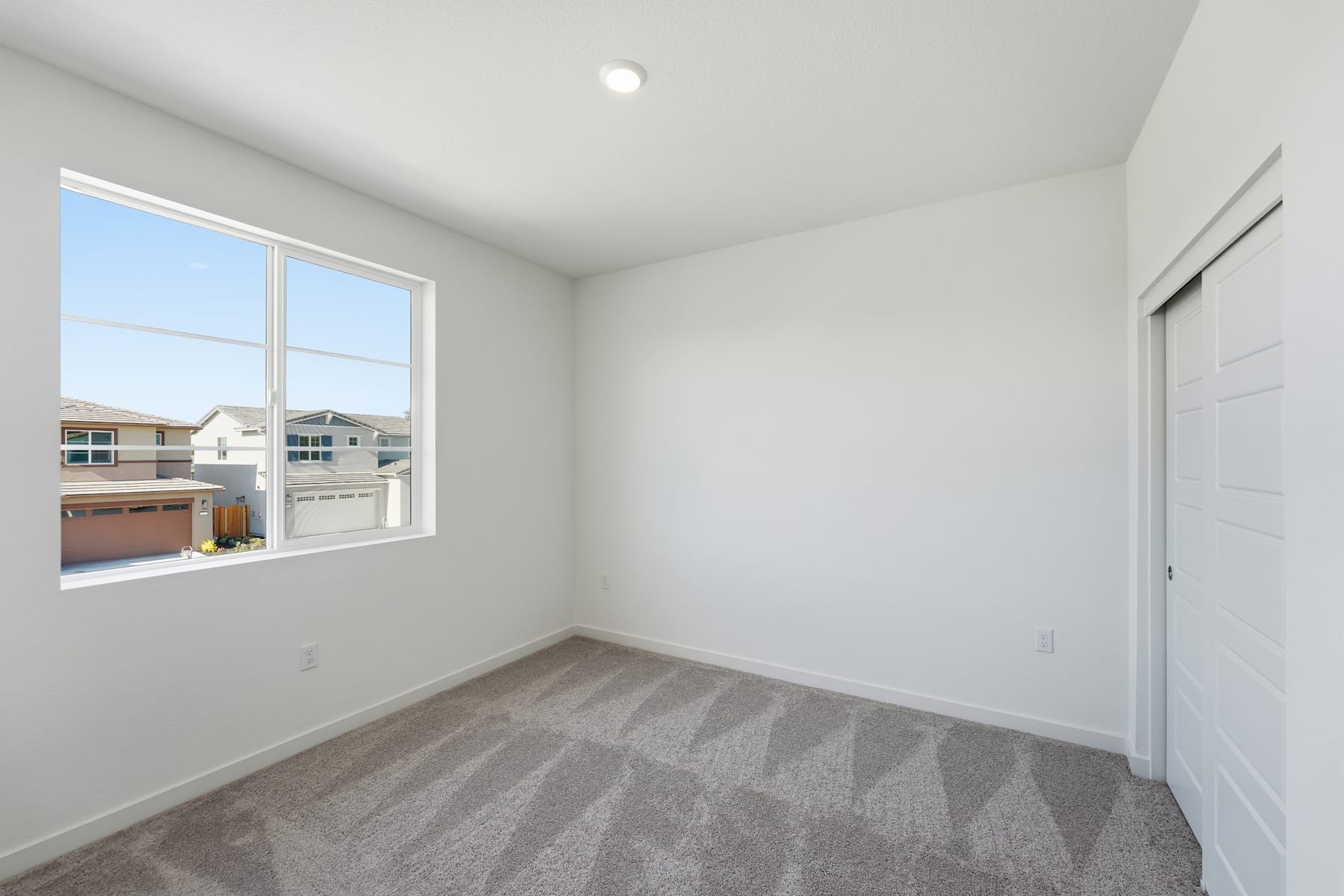 A bright, spacious room with a large window overlooking a residential neighborhood, featuring a gray patterned carpet and a closed door.