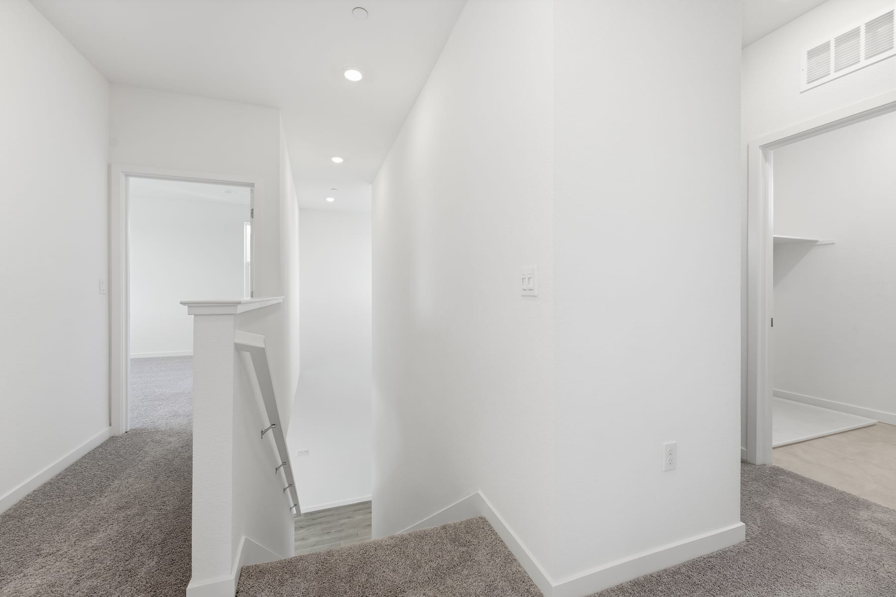 A bright, minimalist hallway with white walls, recessed lighting, and a carpeted floor leading to an open doorway.