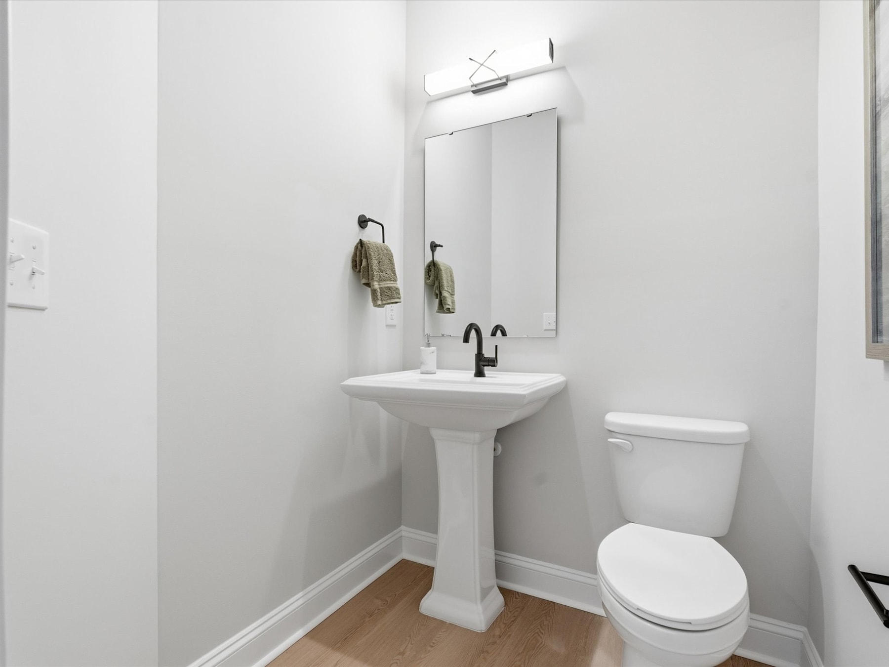 A simple and clean bathroom with a pedestal sink, toilet, and a mirror with a light fixture above it, set against a white wall.
