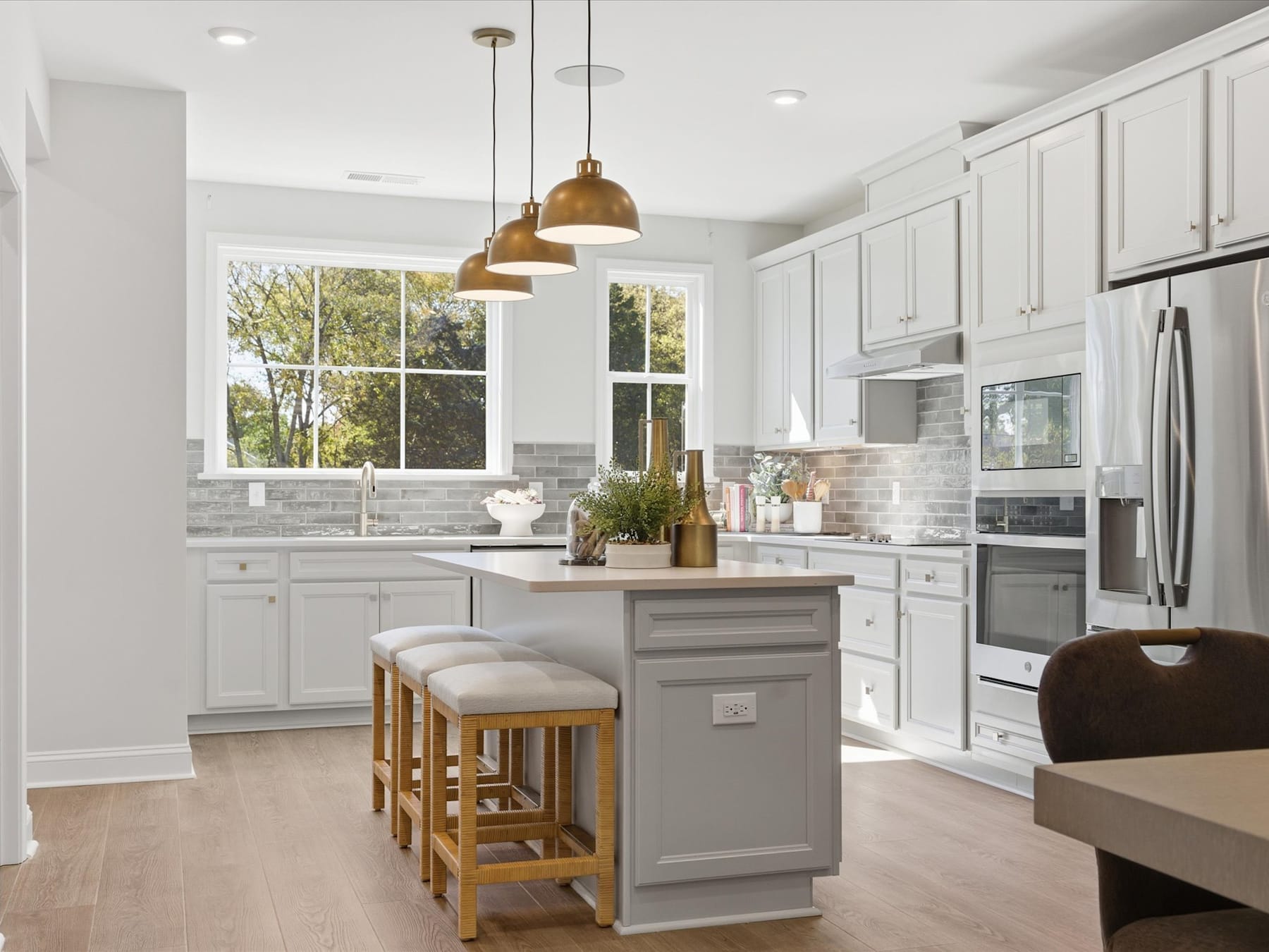 A modern, bright kitchen with white cabinets, a central island, and pendant lighting fixtures, set against a backdrop of large windows overlooking a natural outdoor scene.