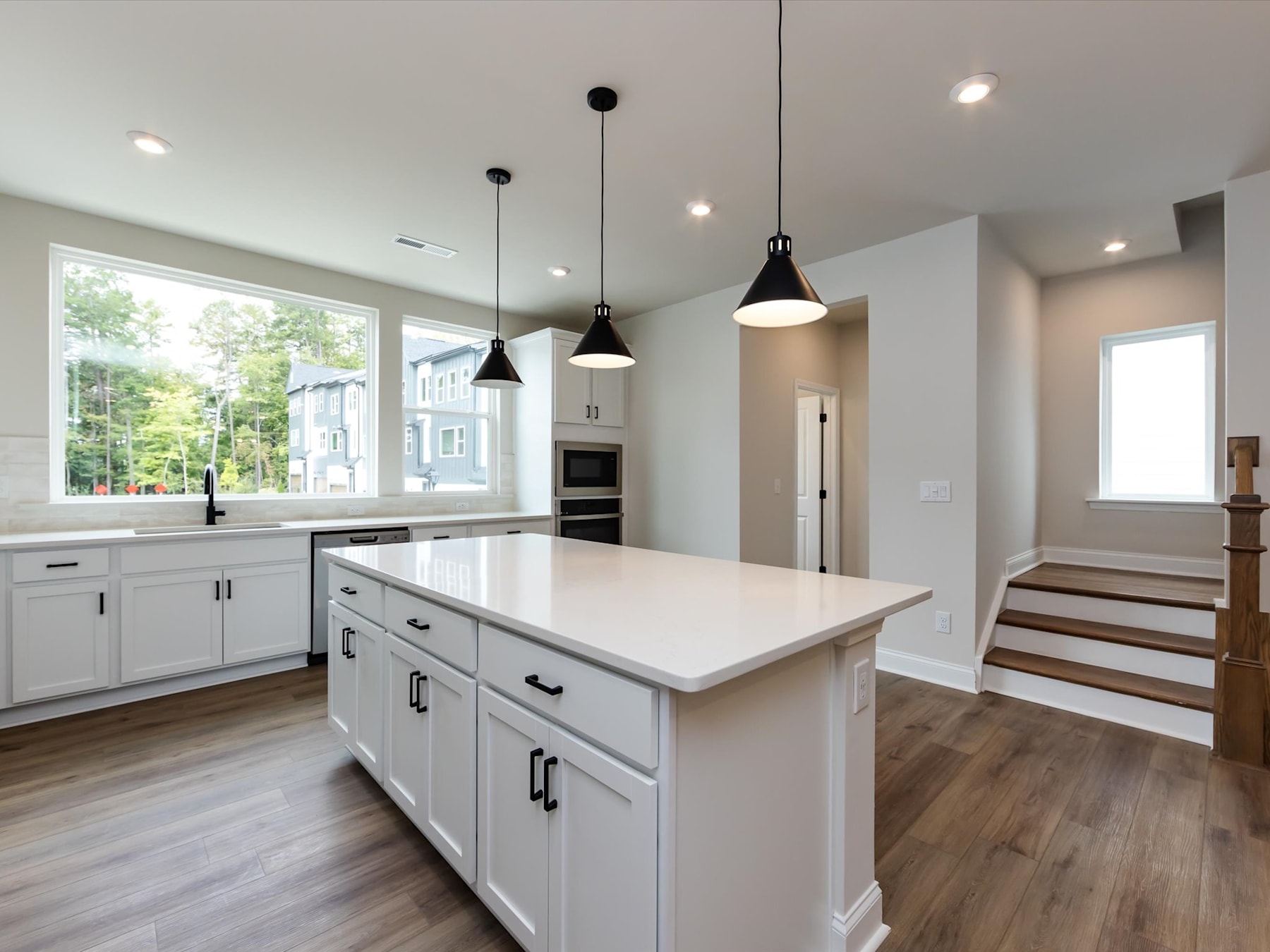 A modern, open-concept kitchen with white cabinets, a large island, and pendant lighting, set against a backdrop of hardwood floors and a staircase leading to another level.