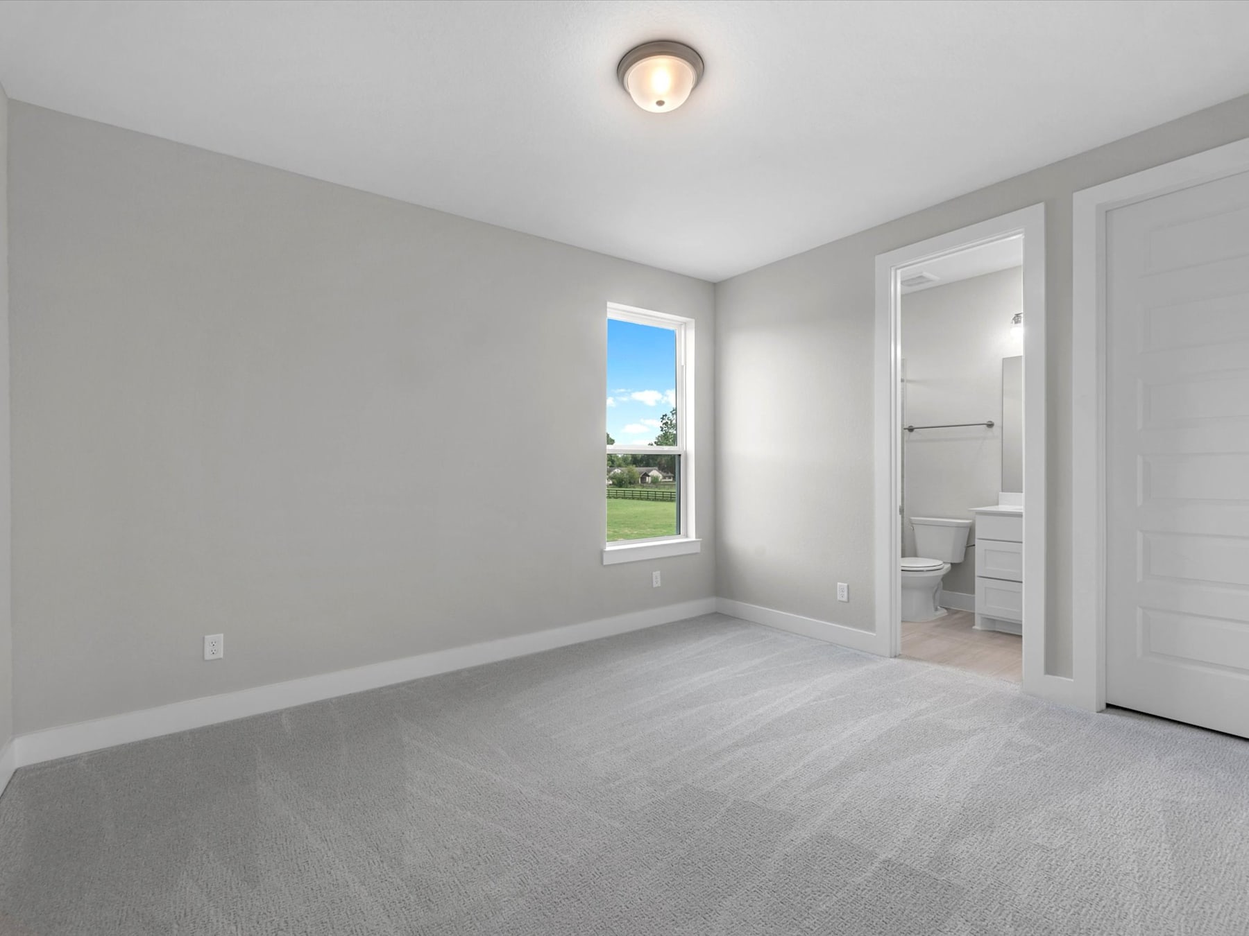 A spacious, minimalist bedroom with a window overlooking a grassy outdoor area, featuring a simple light fixture and a doorway leading to an adjacent room.