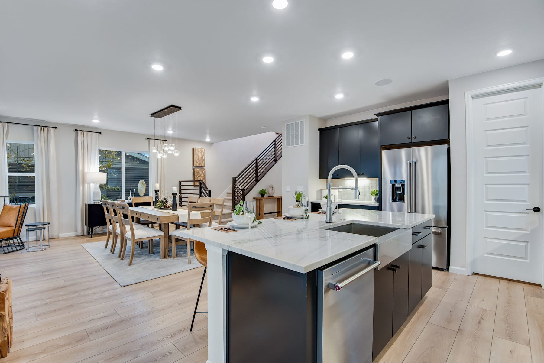A modern, open-concept kitchen and dining area with sleek black cabinetry, stainless steel appliances, and a large central island with a white countertop, surrounded by a spacious living space with hardwood floors and recessed lighting.