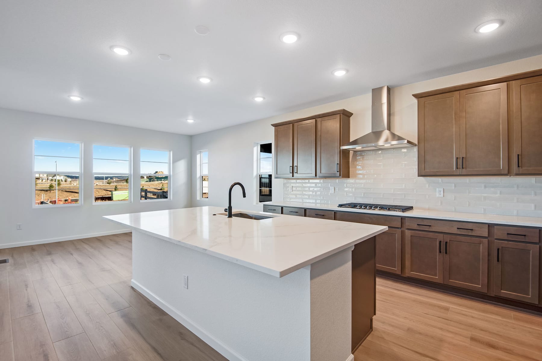 A modern, open-concept kitchen with white countertops, wooden cabinets, and a large island in the foreground, surrounded by large windows offering a scenic view in the background.