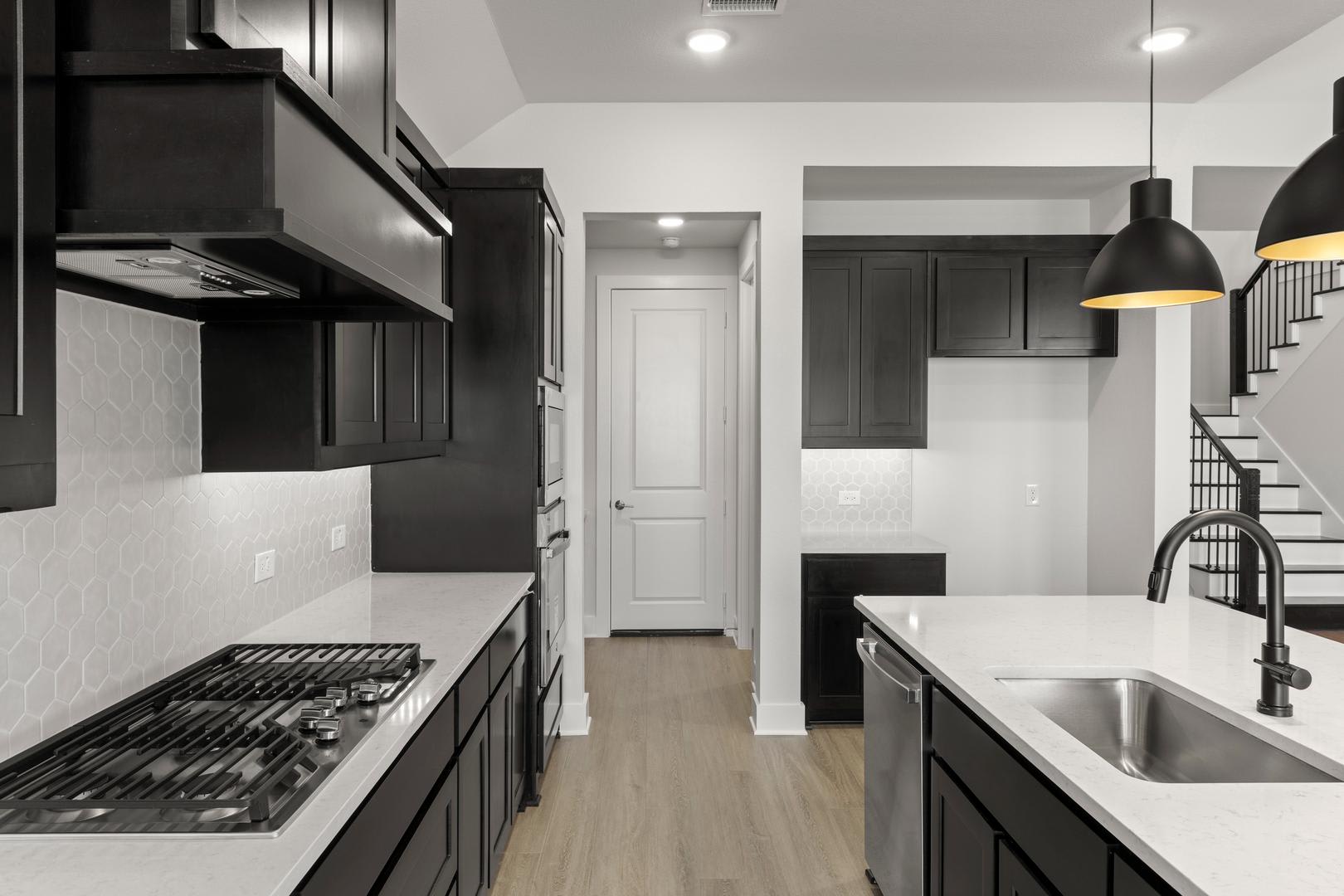 A modern, minimalist kitchen with dark cabinets, a white countertop, and a stainless steel appliance in the foreground, set against a bright, open space in the background.