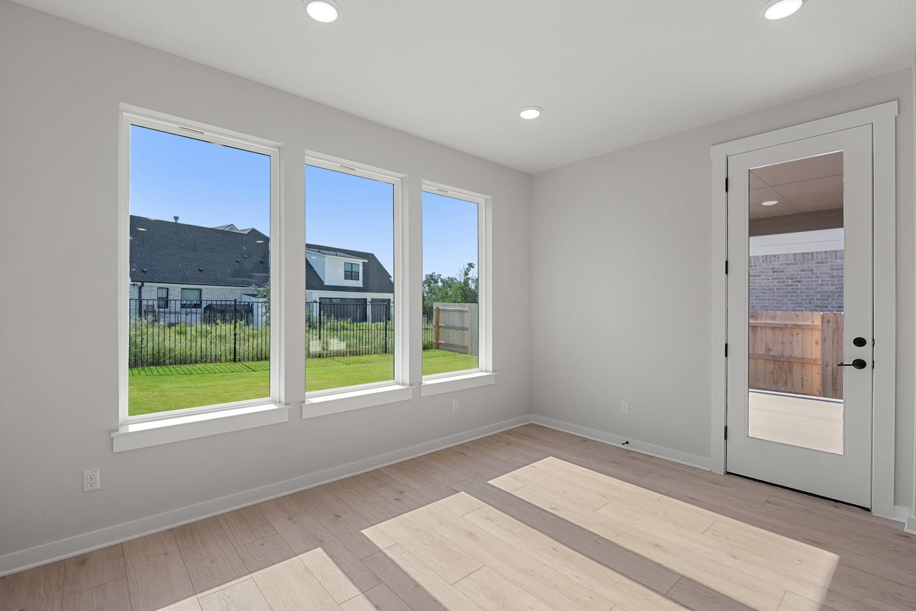 A bright and airy room with large windows overlooking a grassy backyard, featuring a wooden floor and a white door leading to the exterior.