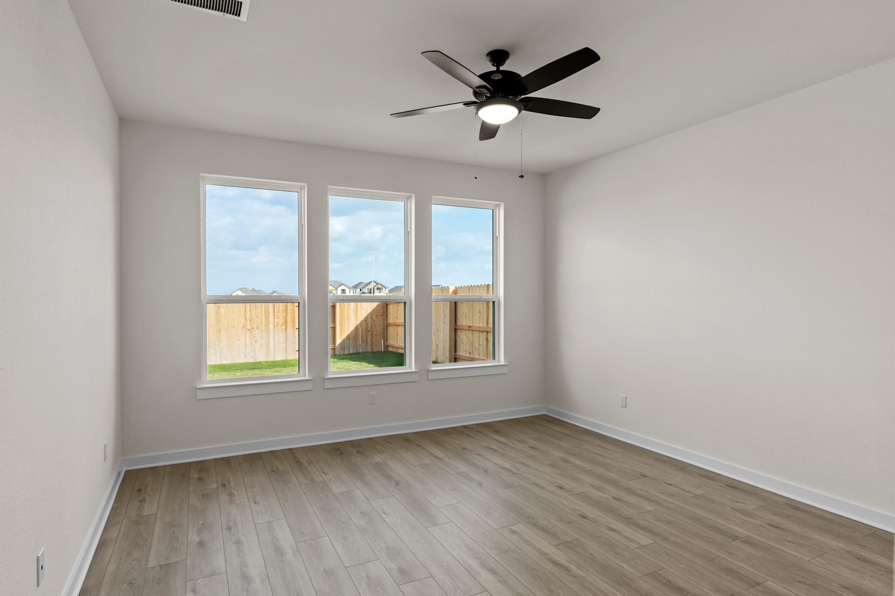 A spacious, well-lit room with a ceiling fan, hardwood-style flooring, and large windows overlooking a fenced backyard.