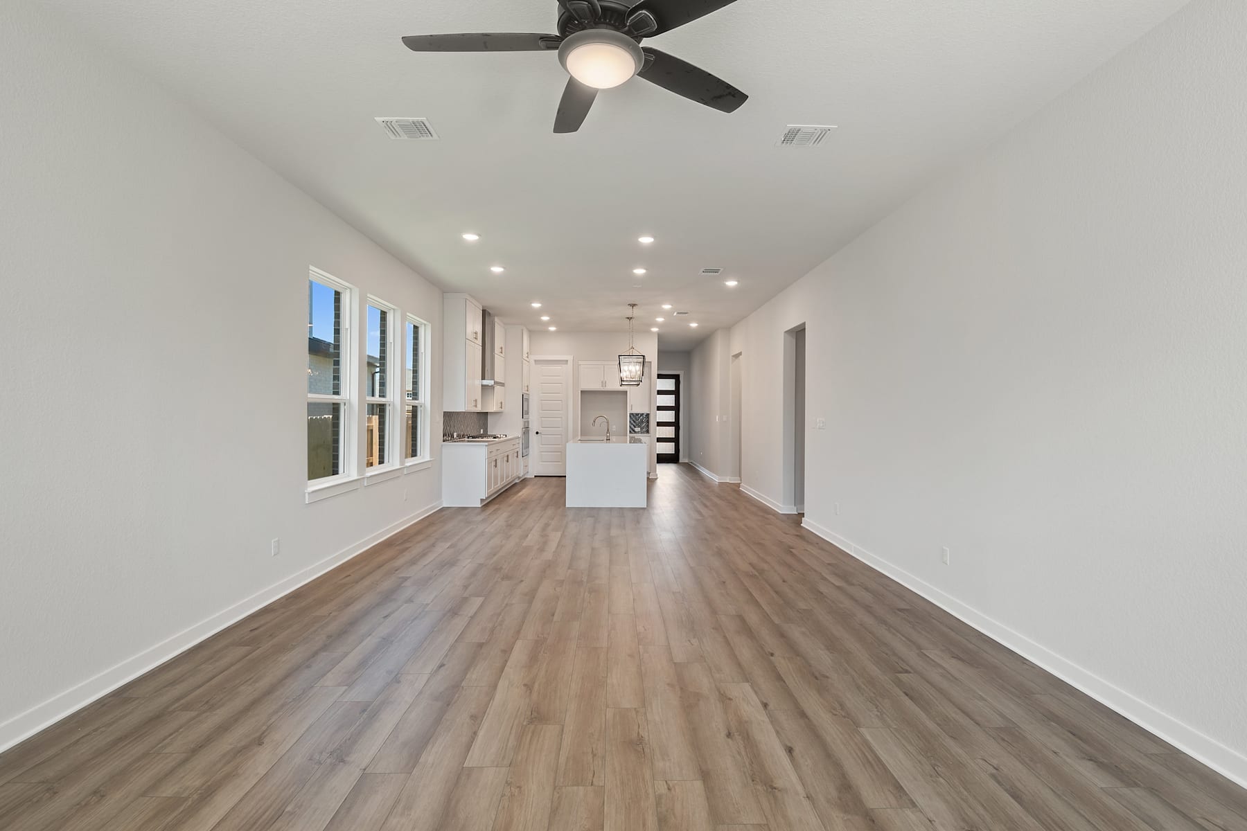 A spacious and well-lit room with a ceiling fan, hardwood floors, and a long hallway leading to a kitchen area.
