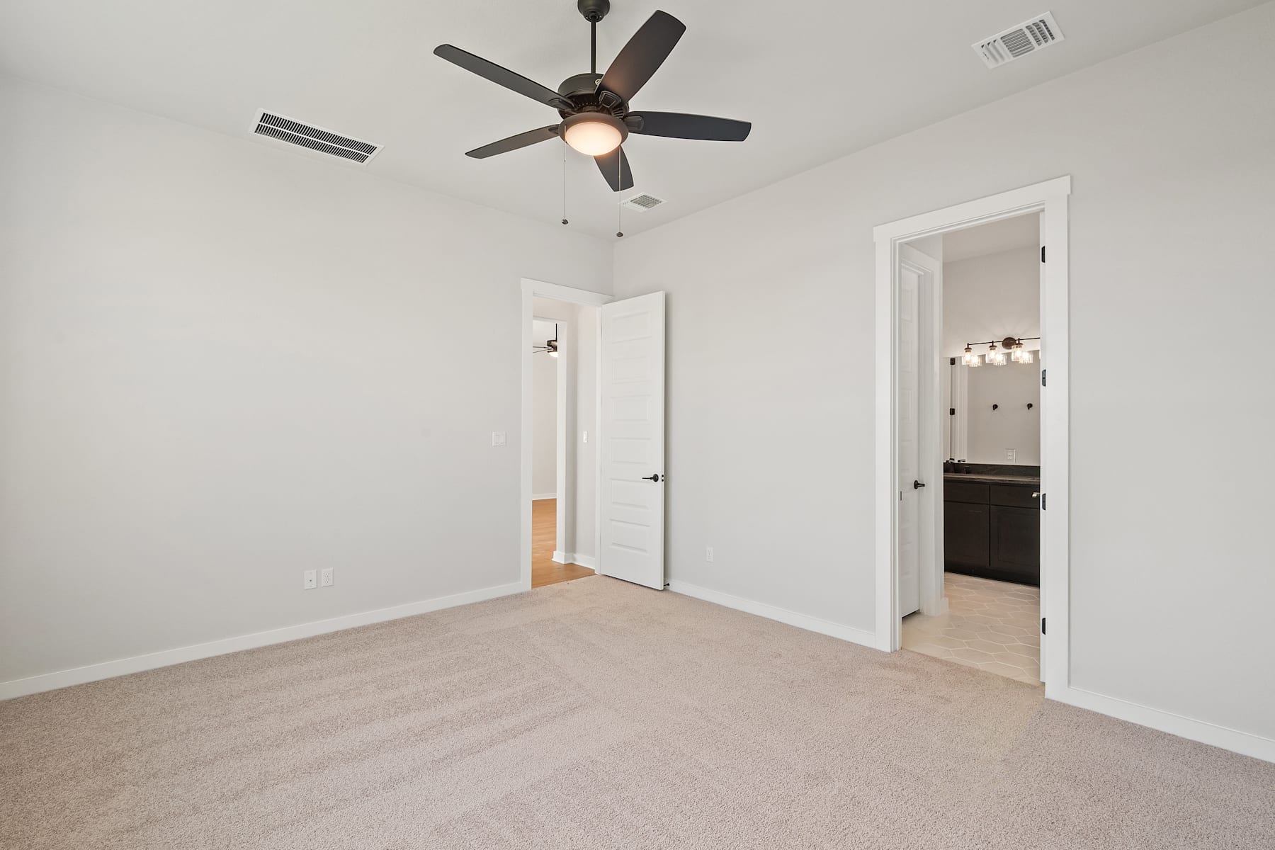 A spacious, well-lit room with a ceiling fan, white walls, and a carpeted floor. The room appears to be empty, with two closed doors visible in the background.