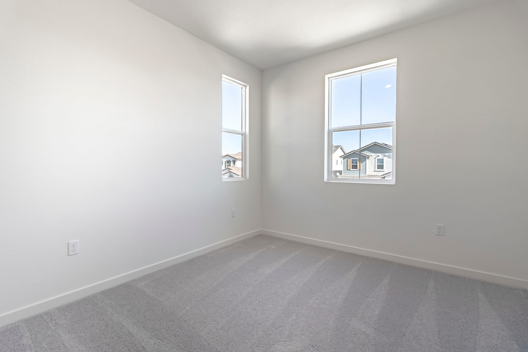 A bright, empty room with white walls and a gray carpeted floor, featuring two windows overlooking residential buildings in the background.