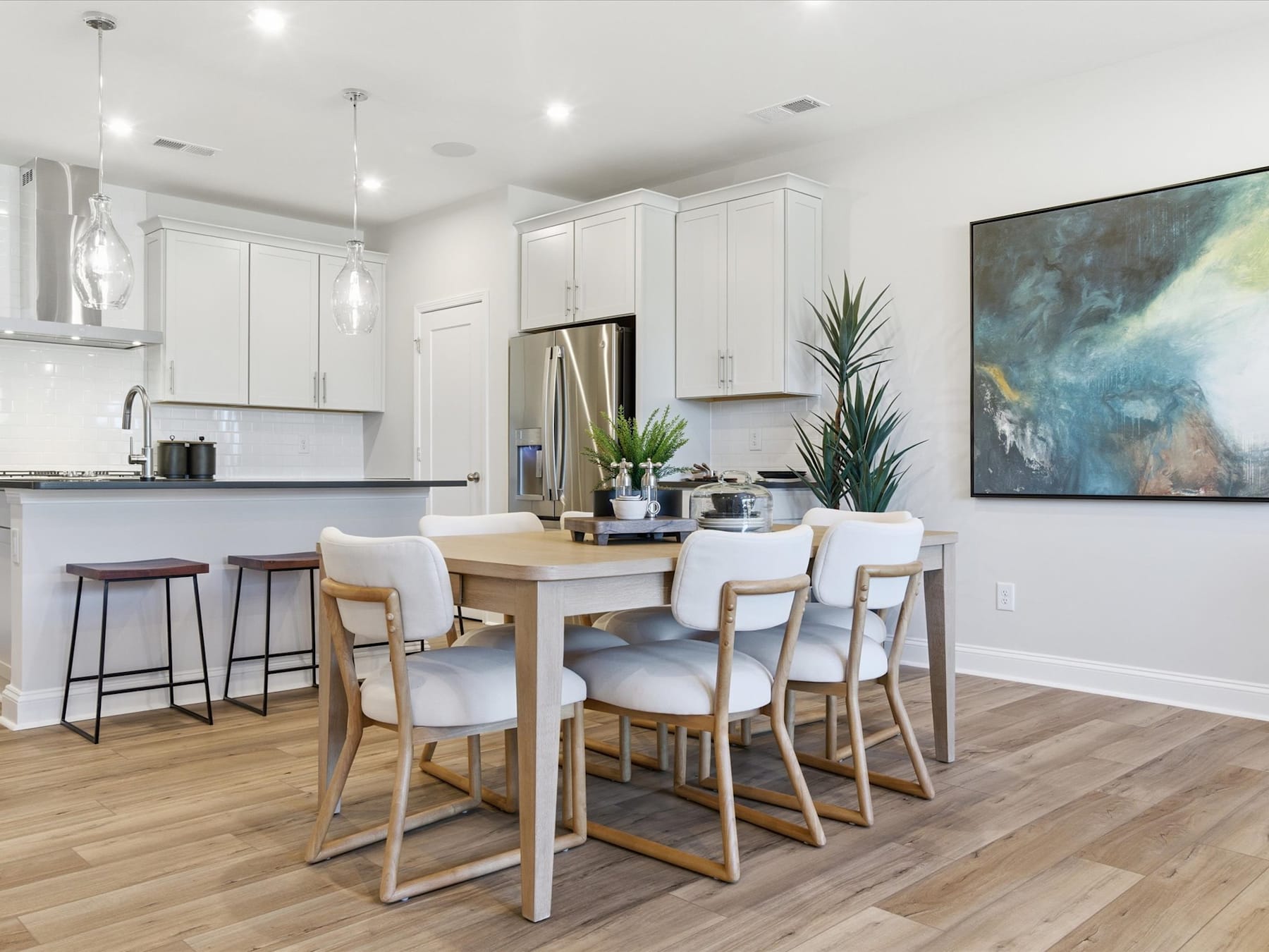 A modern and bright kitchen and dining area with white cabinets, wooden furniture, and a large abstract painting on the wall.