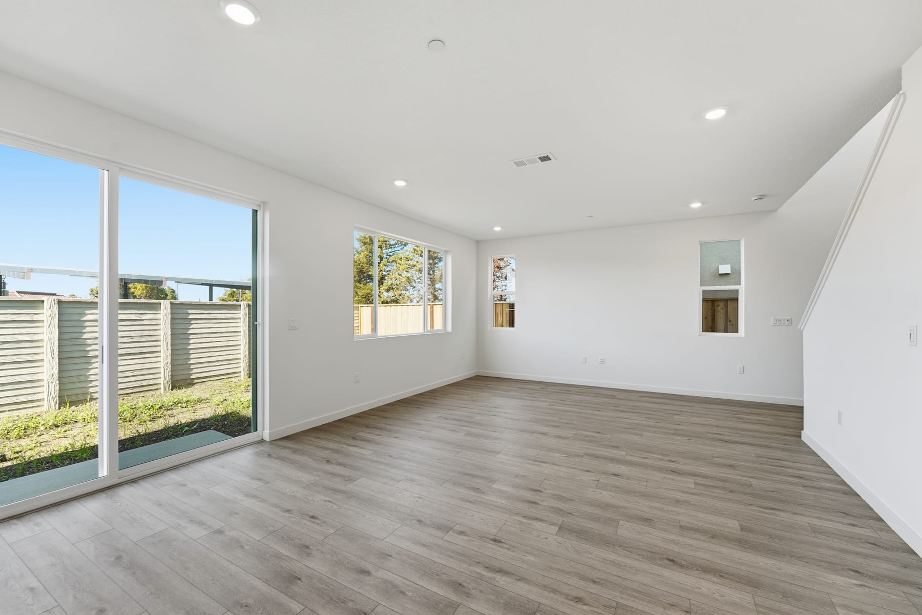 A spacious, well-lit room with large windows overlooking a fenced outdoor area, featuring a hardwood floor and recessed lighting.