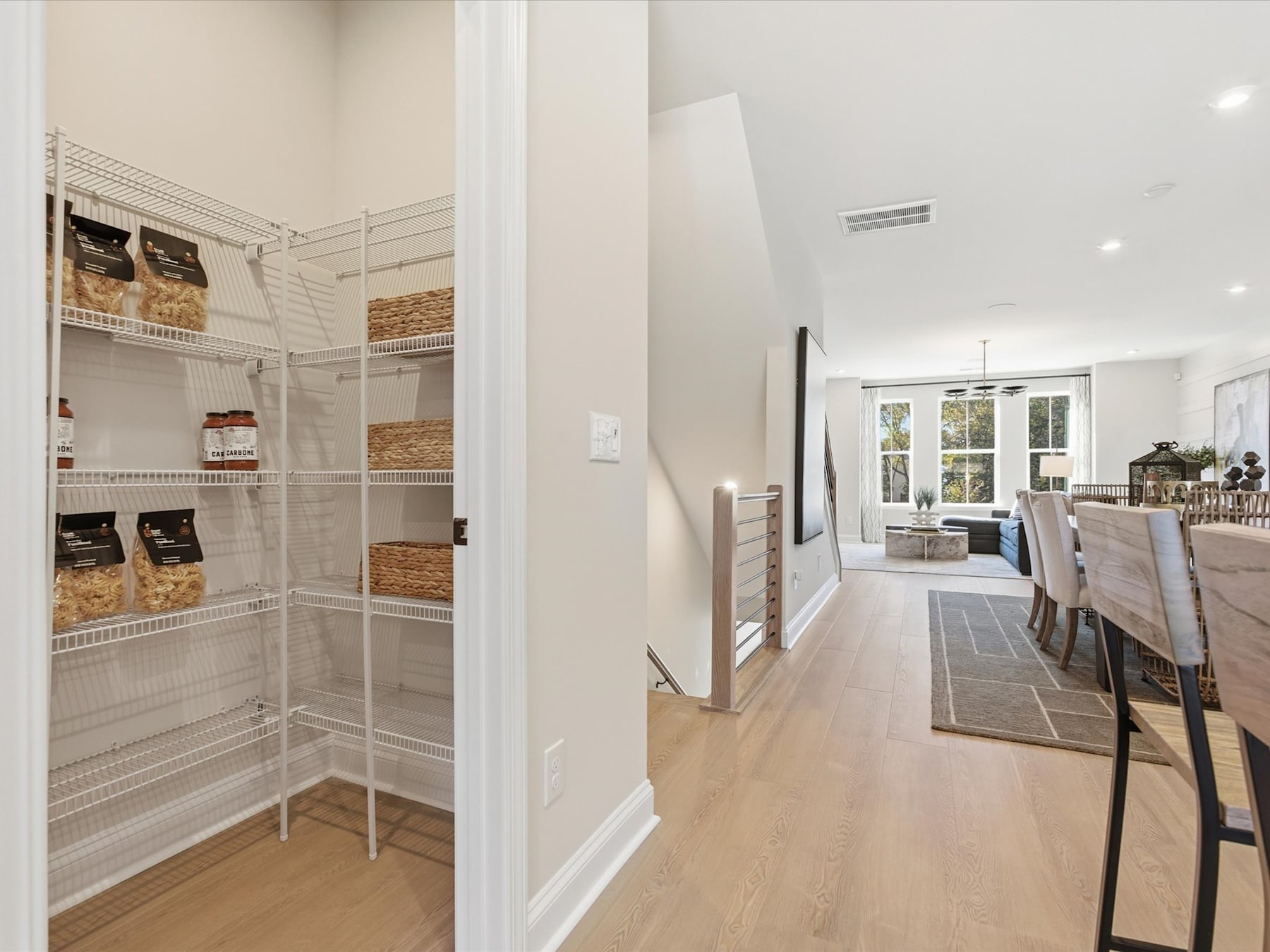 A well-organized pantry with shelves filled with various food items, leading into a bright and open living space with hardwood floors and large windows.